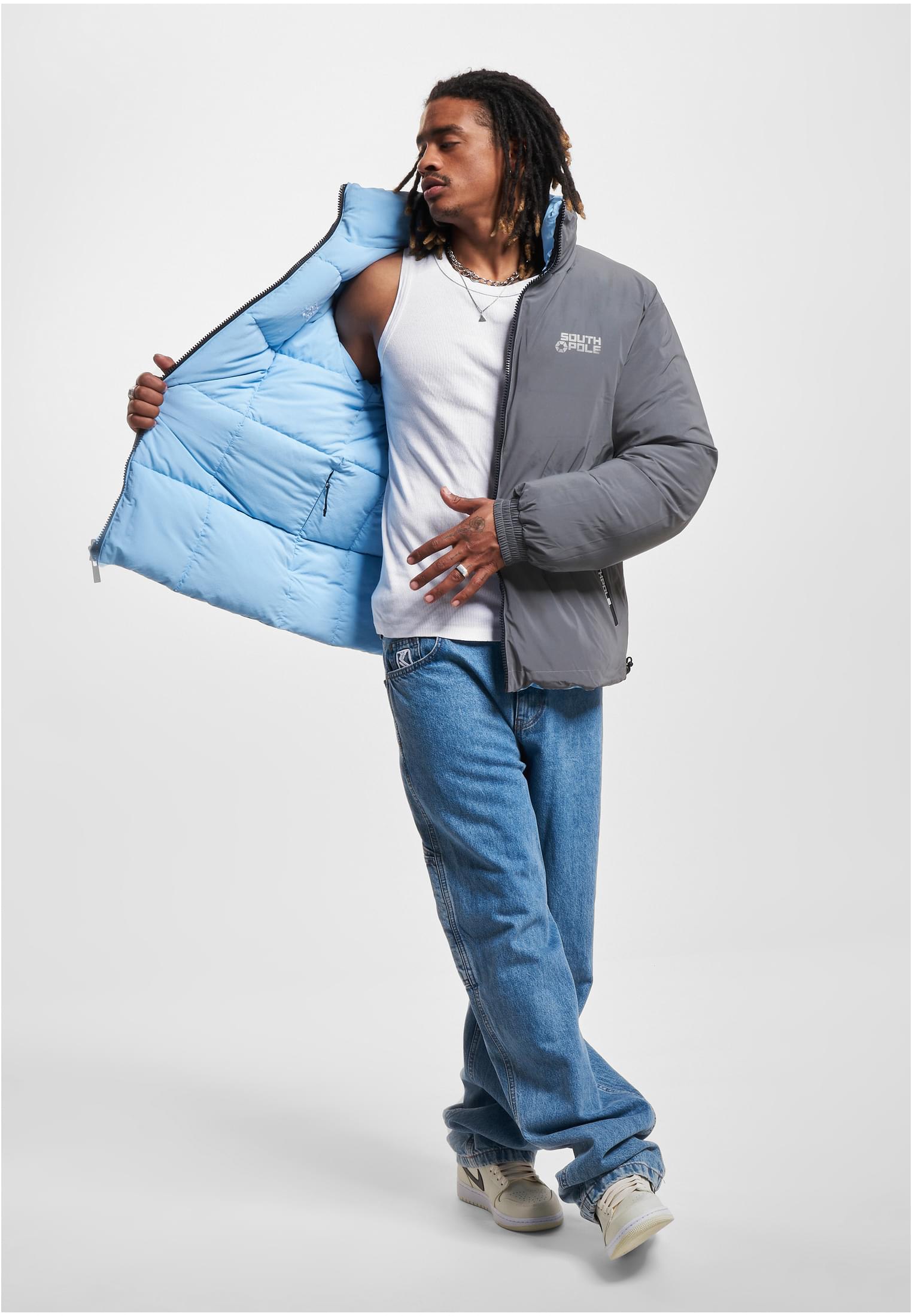 En man i blå jeans och vitt linne öppnar sin gråa Southpole-pufferjacka för att visa det ljusblå fodret.
