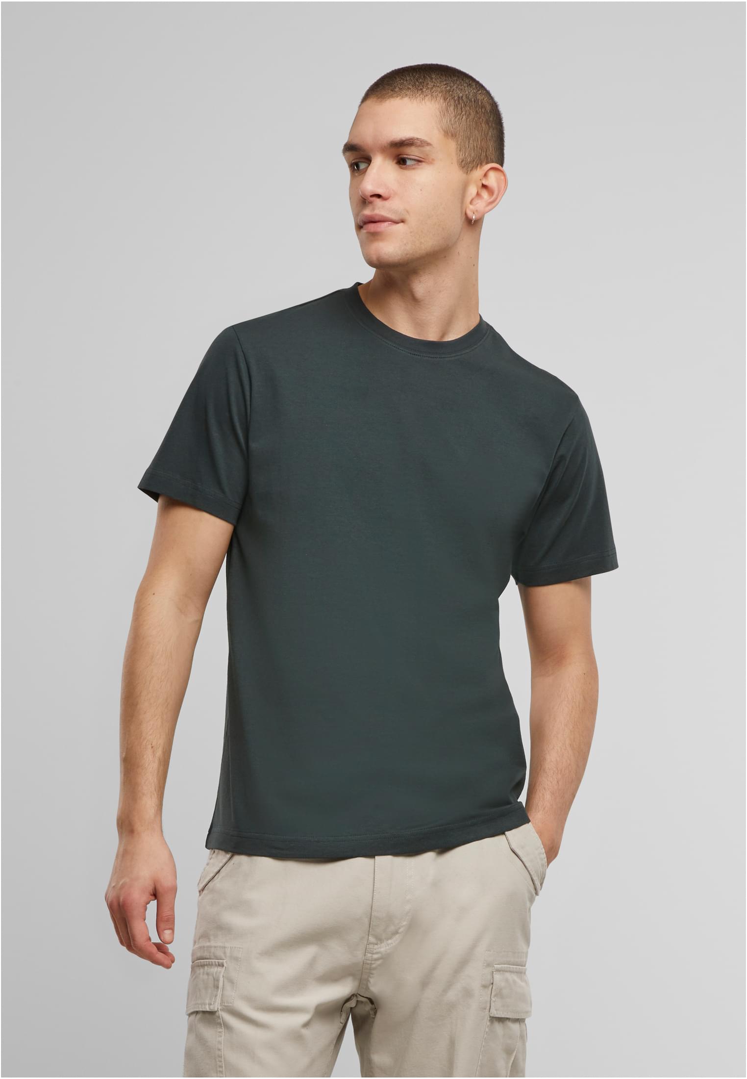 En manlig modell bär en mörkgrön Brandit Premium T-shirt och ljusa cargobyxor. Han står framför en ljusgrå bakgrund och tittar åt sidan.