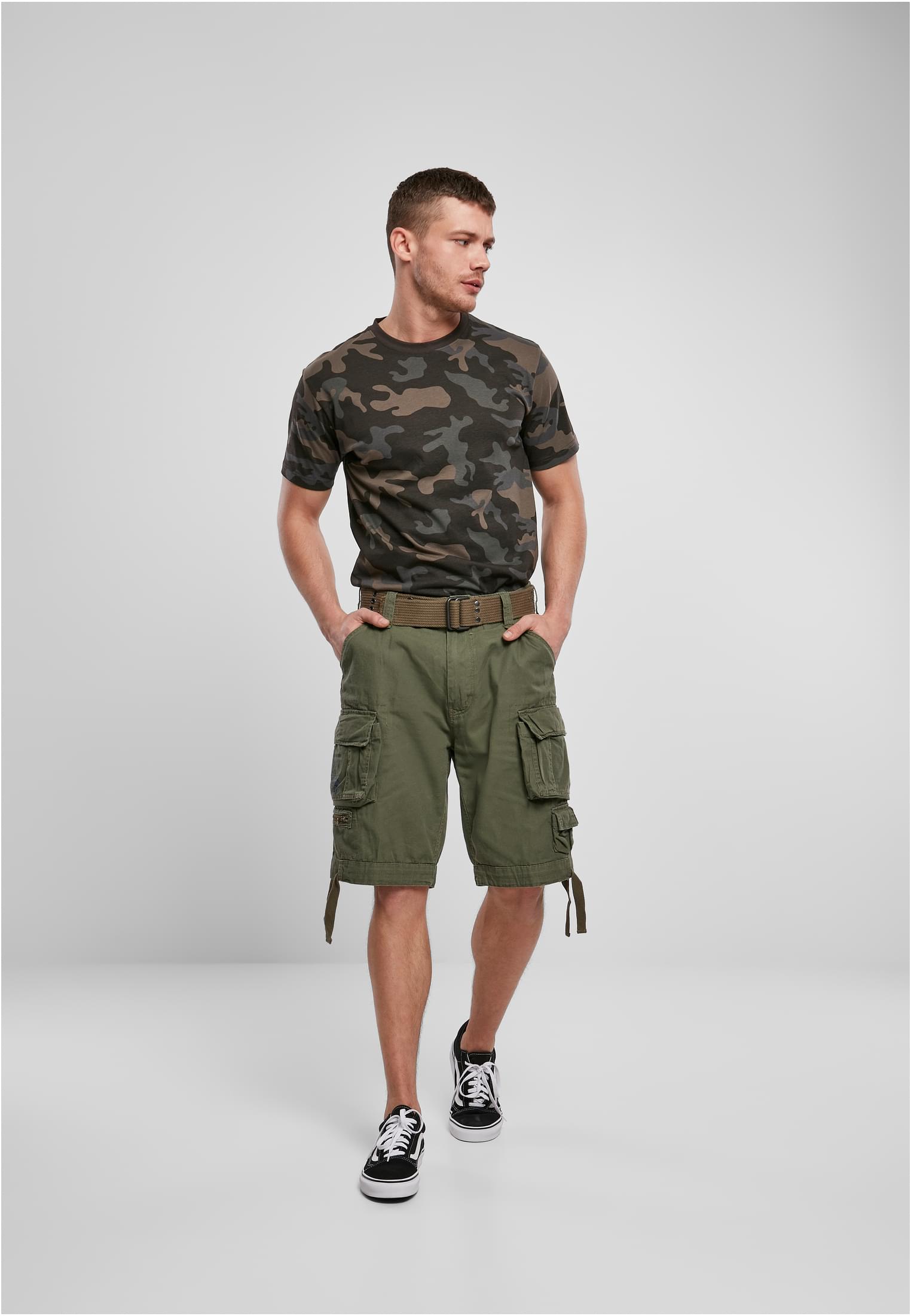 Manlig modell som poserar i en kamouflagemönstrad t-shirt, gröna cargoshorts och svarta sneakers mot en vit bakgrund.