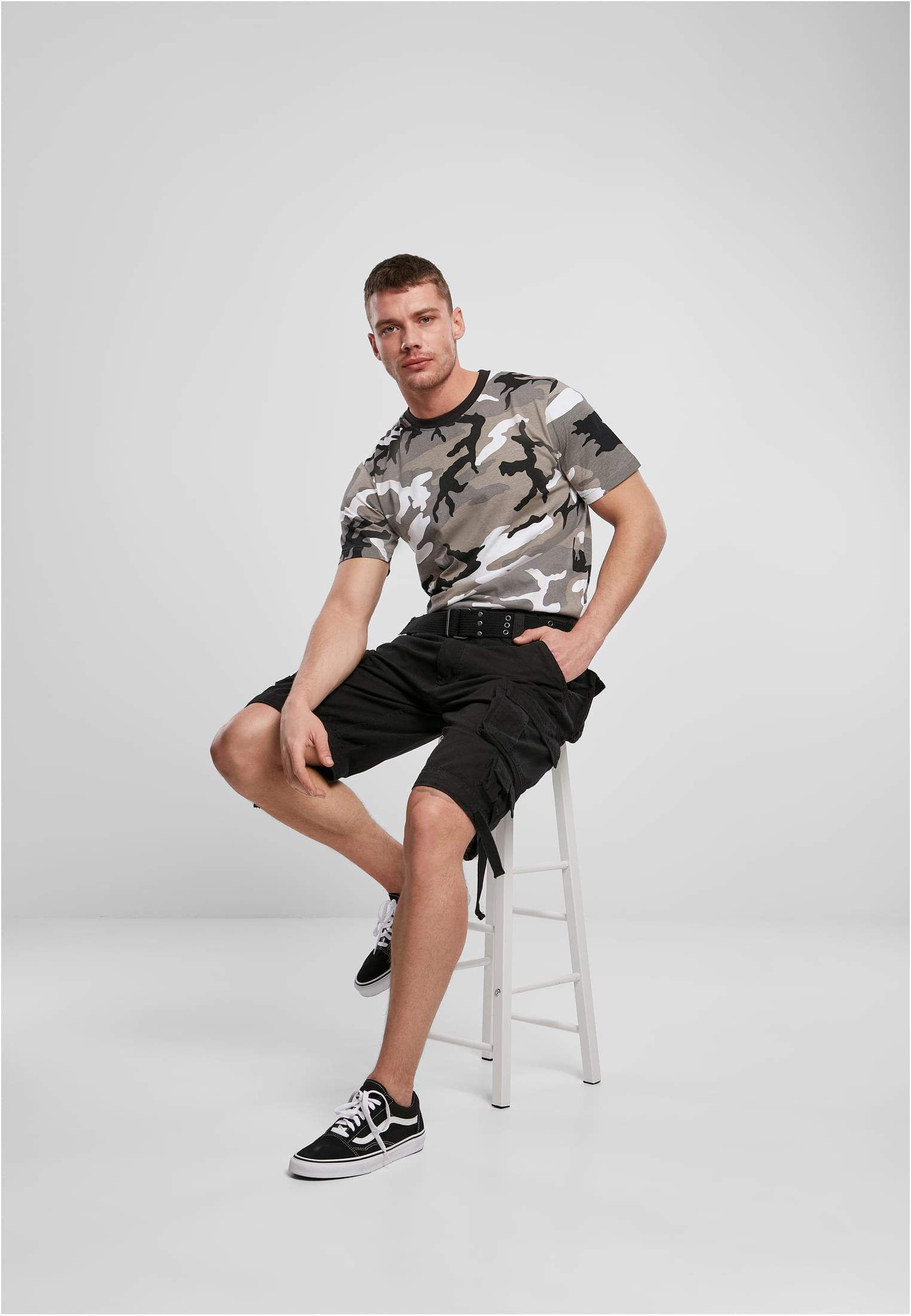 En man sitter på en vit pall och bär en kamouflagemönstrad B Premium Shirt.