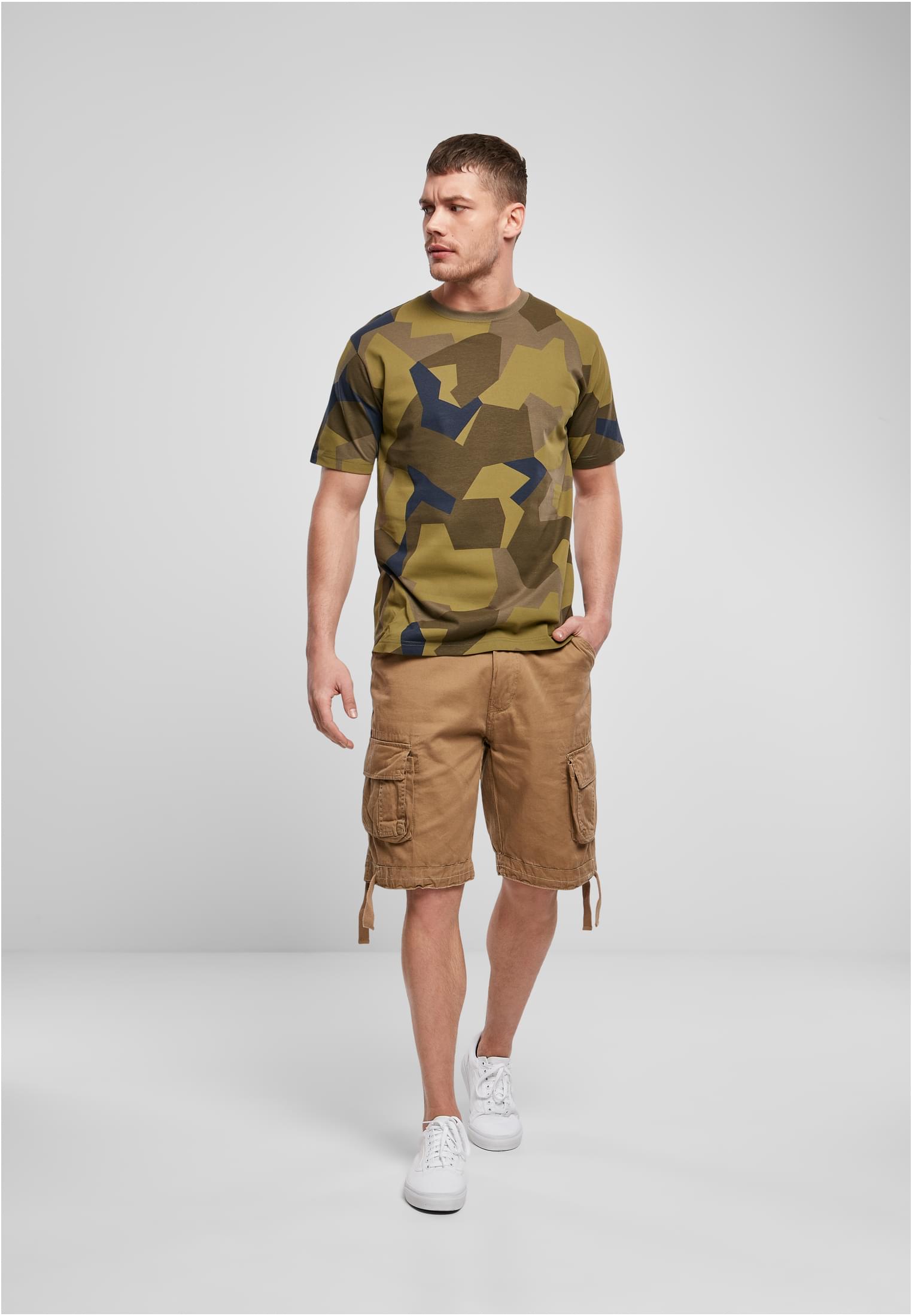Framsida av en manlig modell som bär B Premium Shirt i ett grönt kamouflagemönster, tillsammans med beiga shorts och vita sneakers.