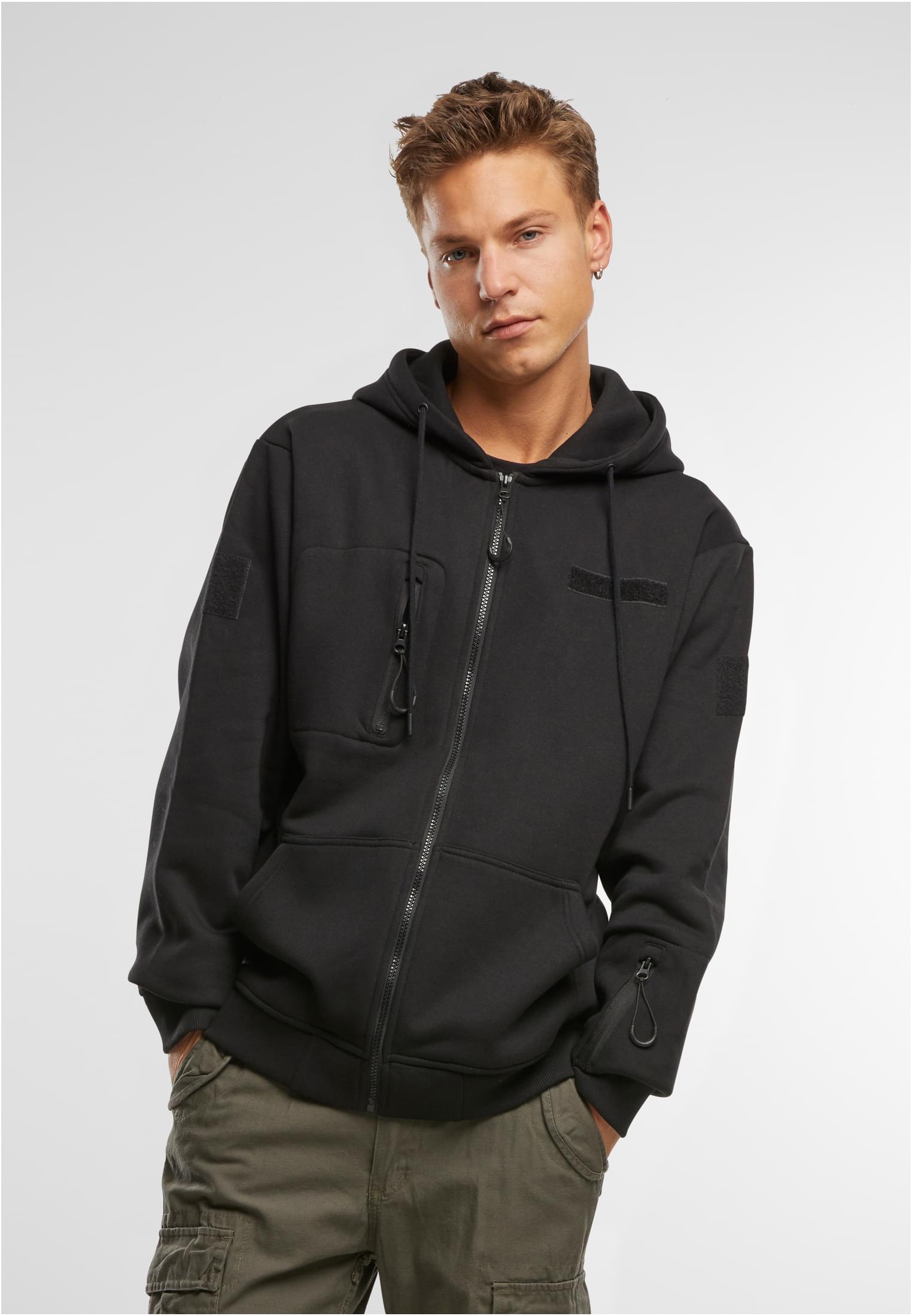 En man poserar i en svart B Tactical Sweat Jacket med händerna i fickorna, fotograferad framifrån mot en vit bakgrund.