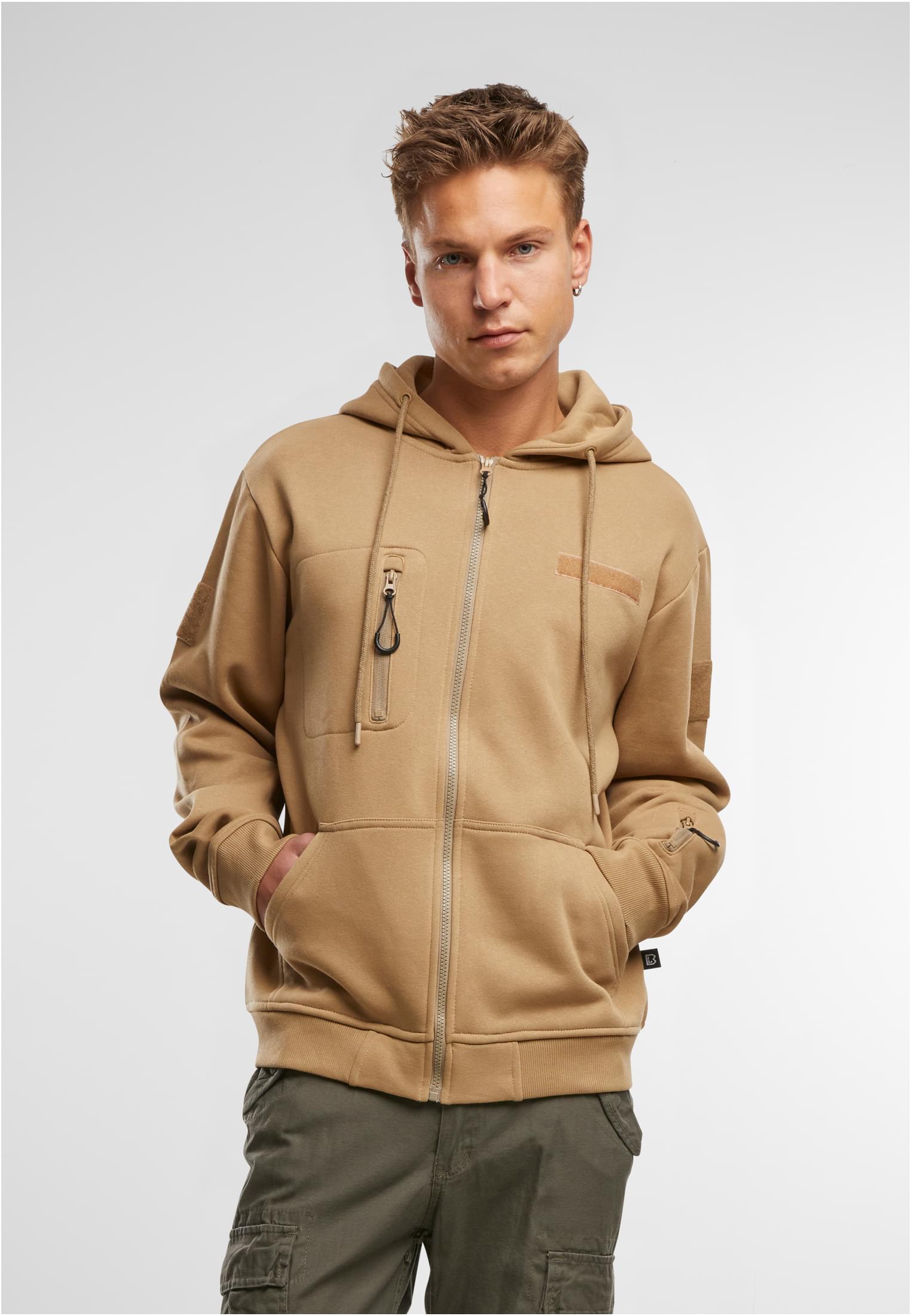 En man visar framsidan av en beige B Tactical Sweat Jacket med händerna i fickorna.