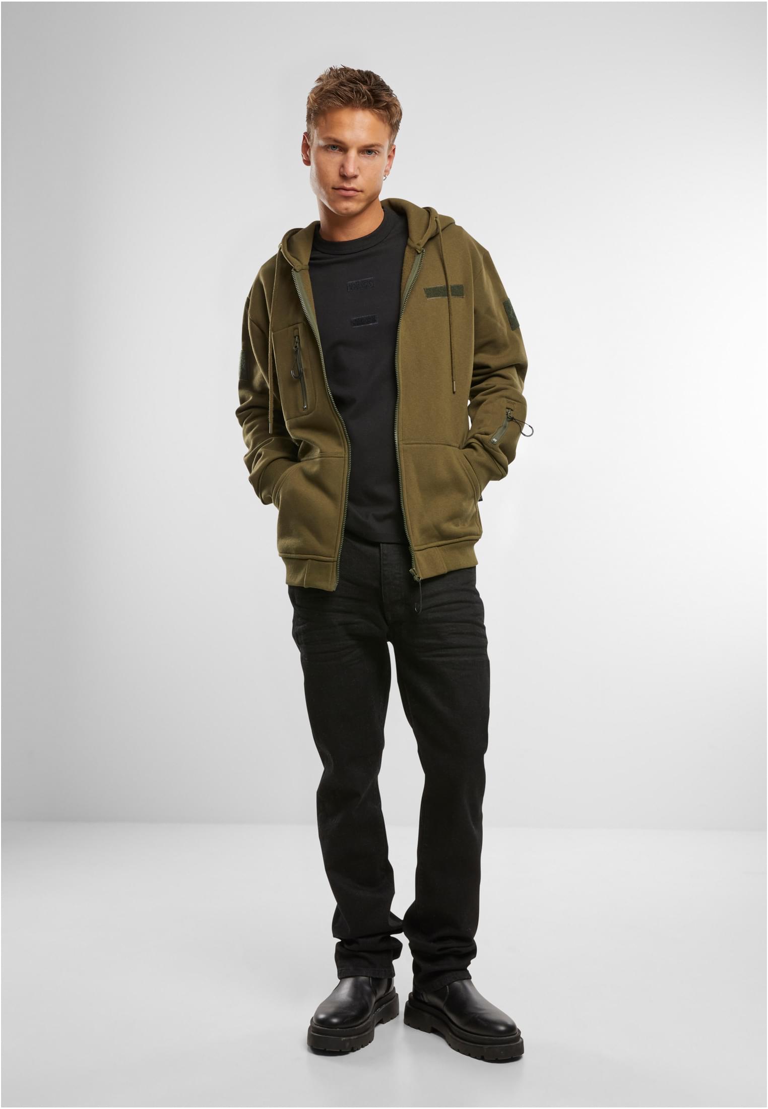 En man visar framsidan av en olivgrön B Tactical Sweat Jacket med dragkedjan öppen, kombinerad med en svart t-shirt och svarta jeans.