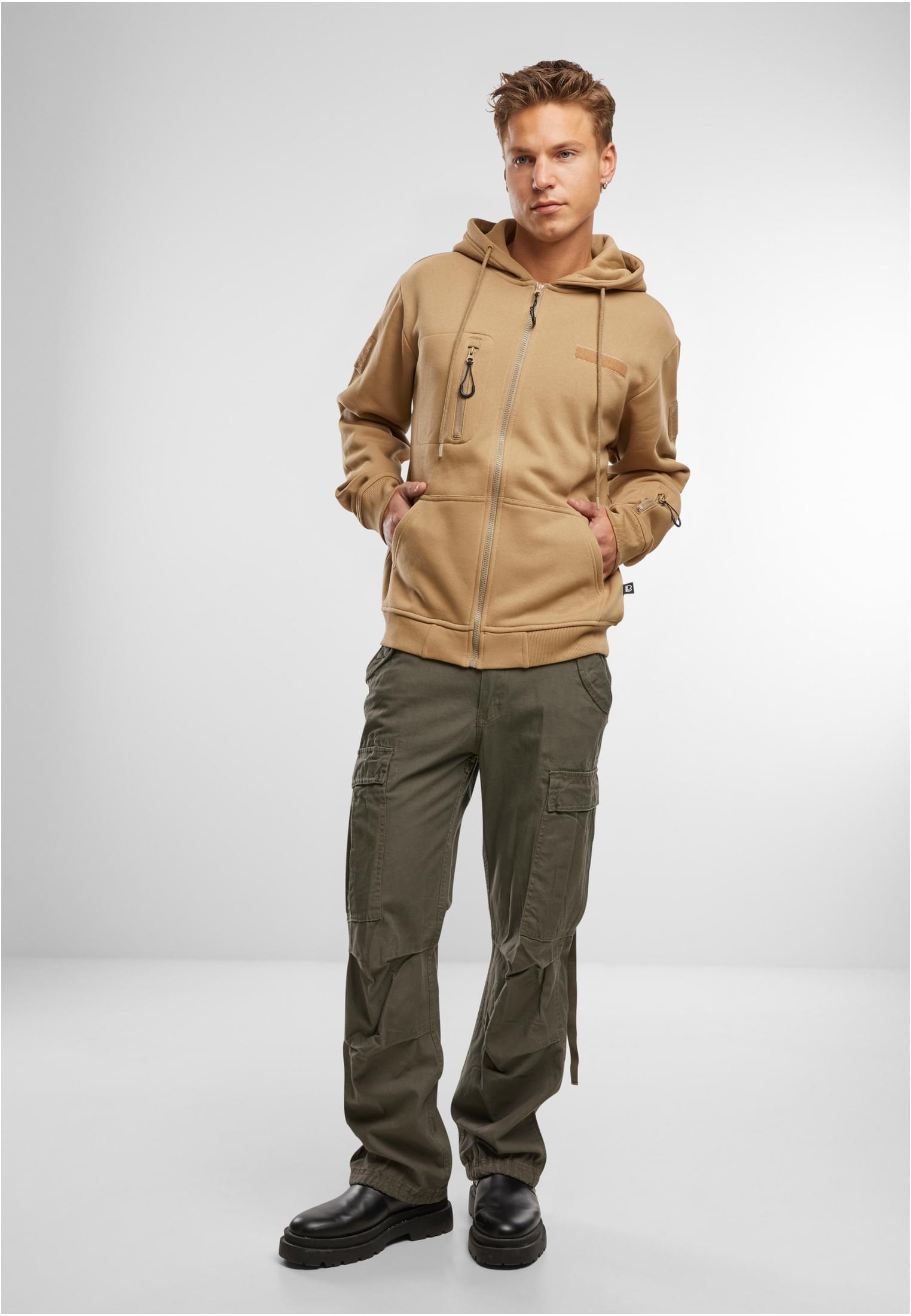 En man poserar i en beige B Tactical Sweat Jacket, olivgröna cargobyxor och svarta kängor mot en vit bakgrund.