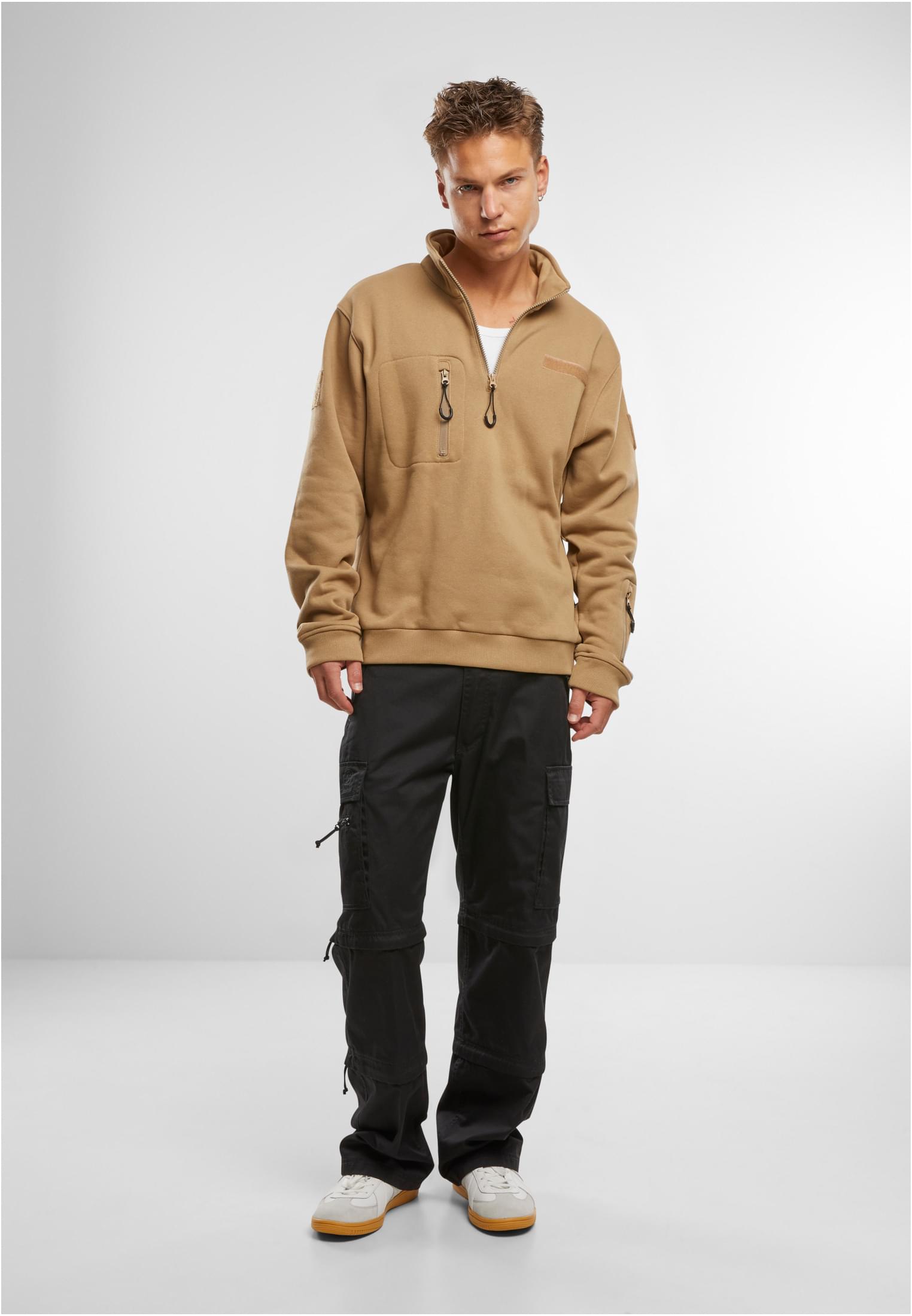 En man visar framsidan av en beige B Tactical Sweat Troyer-tröja, stylad med svarta cargobyxor och vita sneakers.