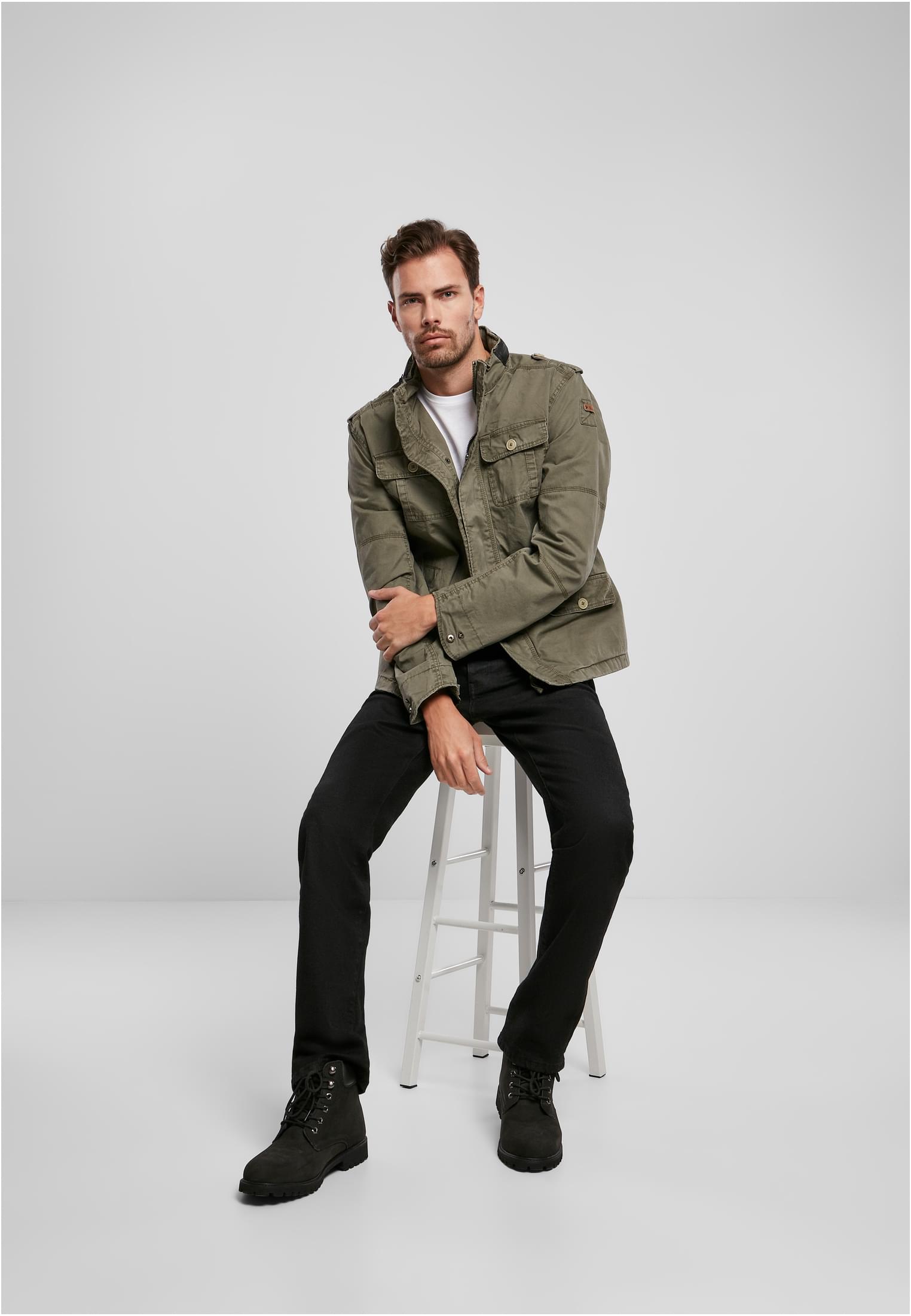 En man visar upp B Britannia-jackan i olivgrönt, stylad med svarta jeans och kängor, sittandes på en pall.