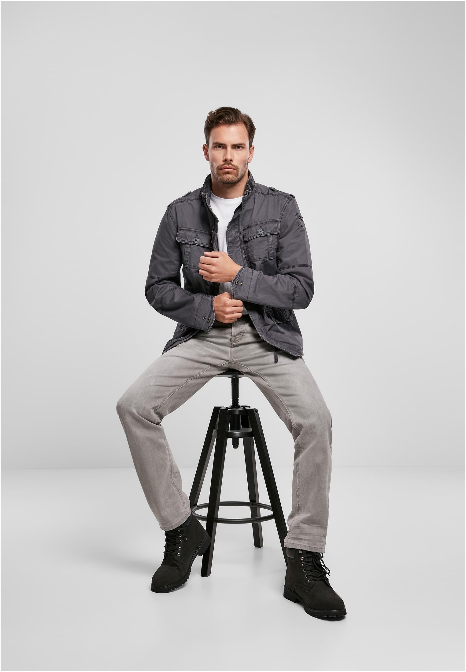 En man sitter på en svart pall, vänd mot kameran, och bär en mörkgrå Britannia-jacka, ljusgrå jeans och svarta kängor.