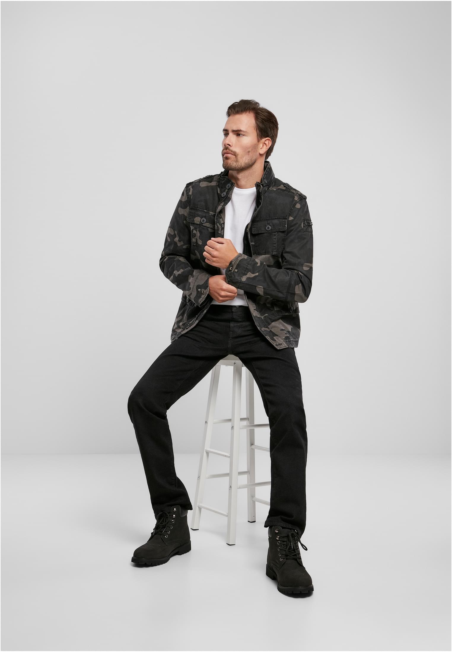 En man sitter på en pall mot en vit bakgrund och bär B Britannia-jackan i svart kamouflagemönster, tillsammans med en vit t-shirt, svarta jeans och svarta kängor.