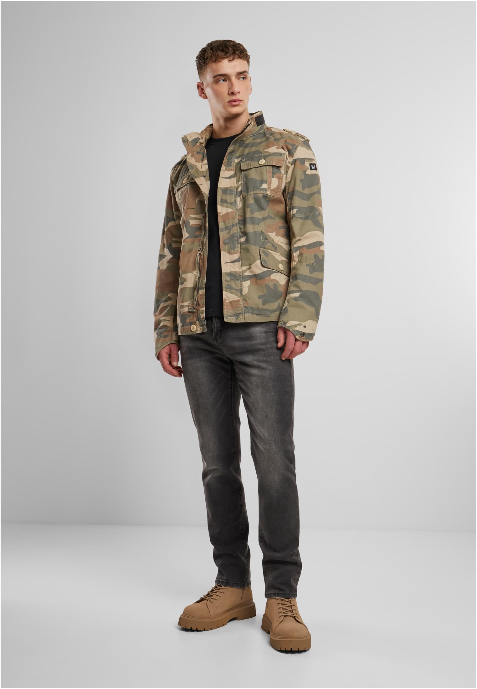 En man visar framsidan av B Britannia Jacket i kamouflagemönster, stylad med en svart t-shirt och mörka jeans.