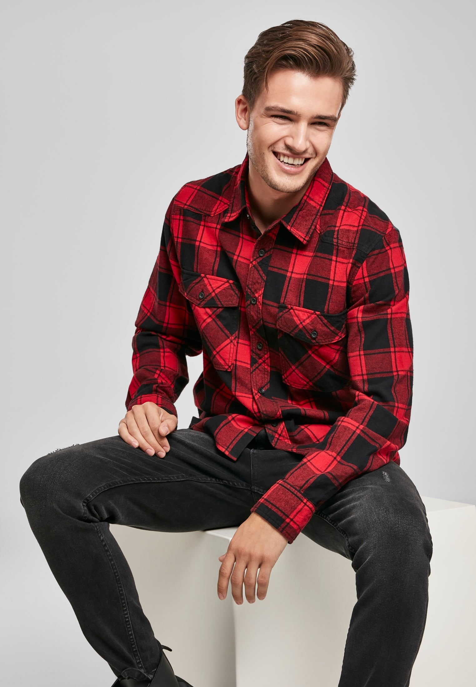 En leende man sitter på en vit kub och bär en röd- och svartrutig B Checkshirt tillsammans med svarta jeans.