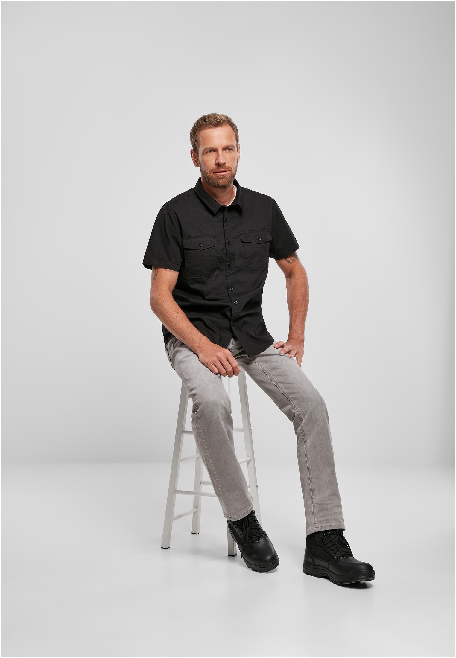 En man sitter på en vit pall och bär en svart kortärmad B Roadstar-skjorta, med gråa jeans och svarta kängor.