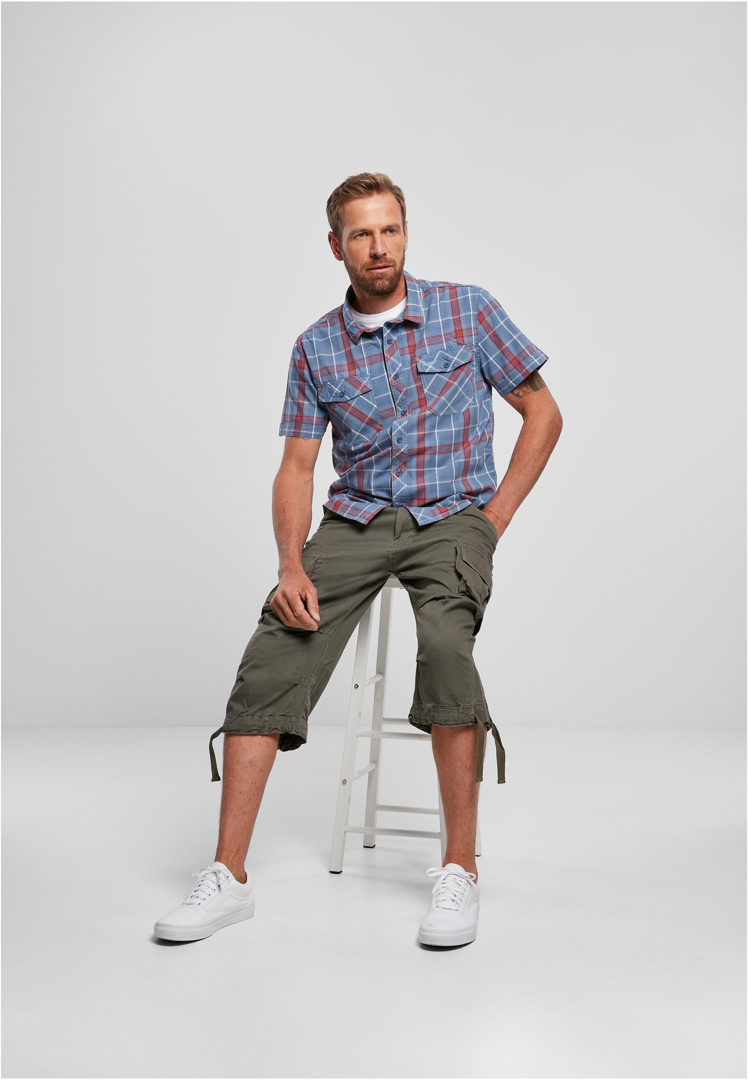 En man sitter på en vit pall och bär en blå och rödrutig kortärmad B Roadstar-skjorta, olivgröna cargoshorts och vita sneakers.