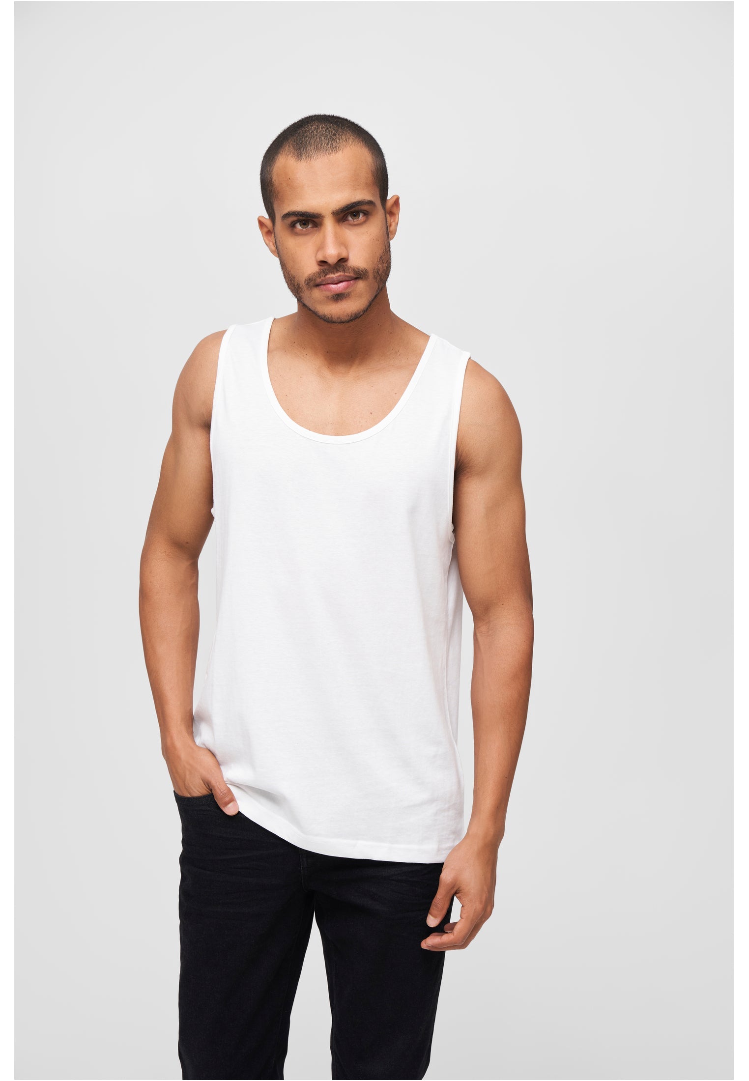 En man i ett vitt B Tank Top-linne och svarta byxor står vänd mot kameran.