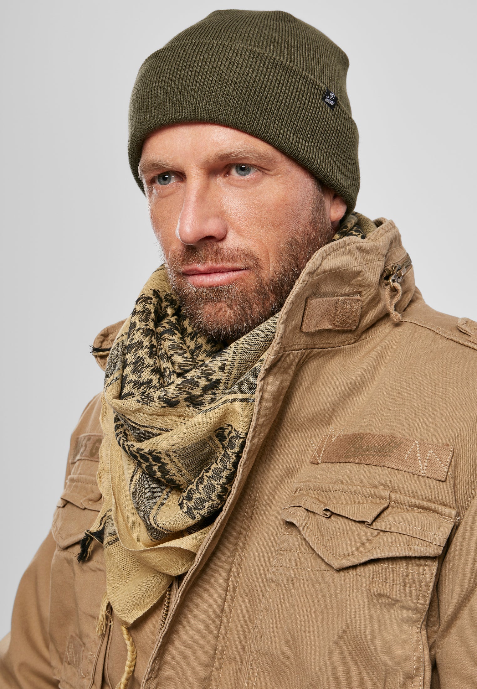En man poserar i en olivgrön B Watch Cap-mössa, en mönstrad scarf och en beige jacka.