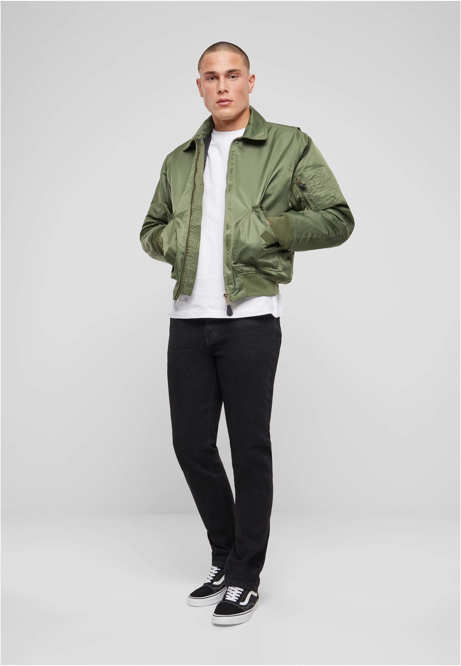 En man visar framsidan av den olivgröna B CWU-jackan från Brandit, buren med en vit t-shirt och svarta jeans.