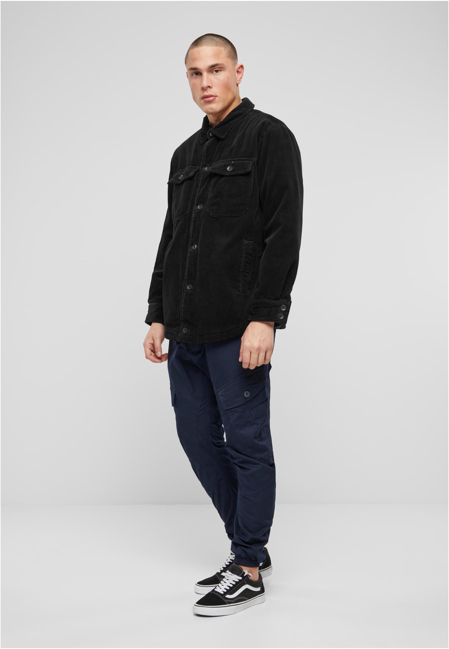 En man poserar i en svart manchesterjacka, mörkblå cargobyxor och svarta sneakers mot en ljusgrå bakgrund.