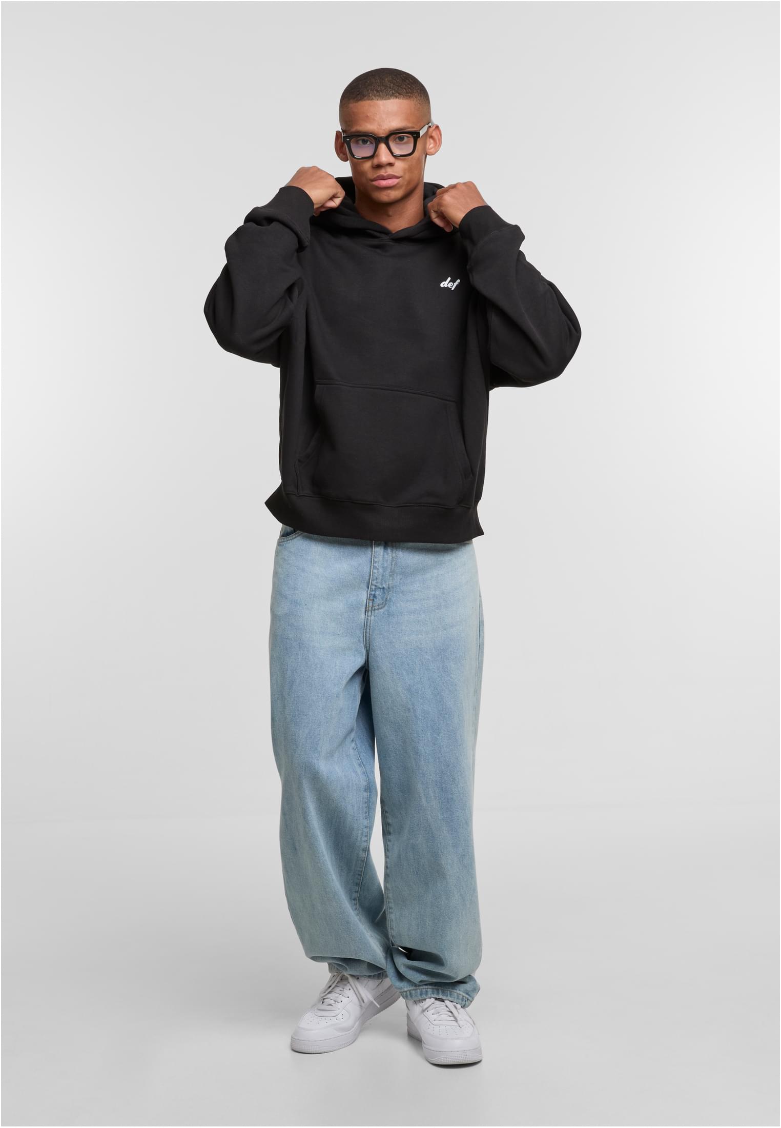En man poserar i den svarta huvtröjan DEF VISIONS med ljusblå jeans och justerar luvan.