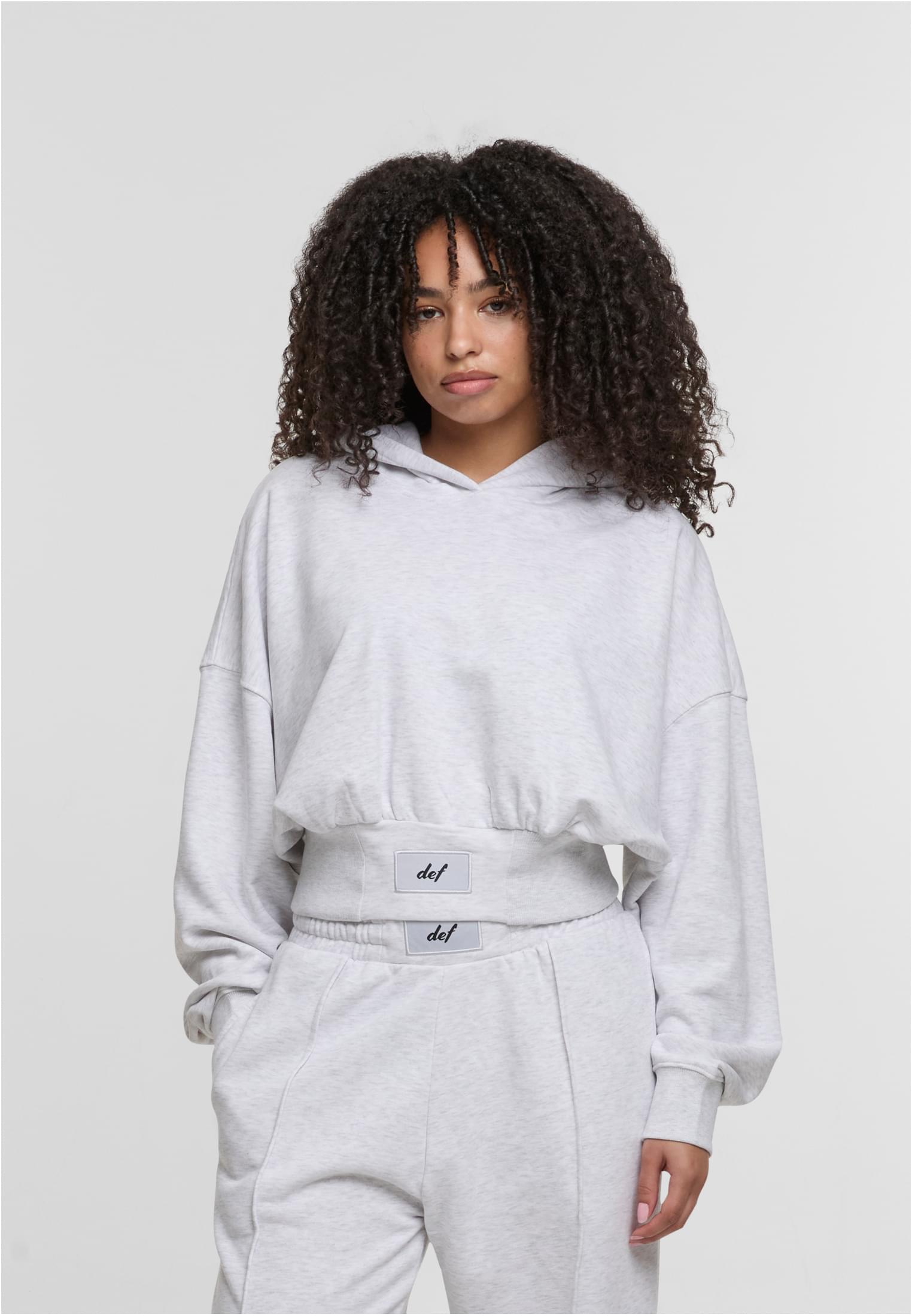 En kvinna bär en ljusgrå, croppad DEF NICE Hoodie och matchande byxor.