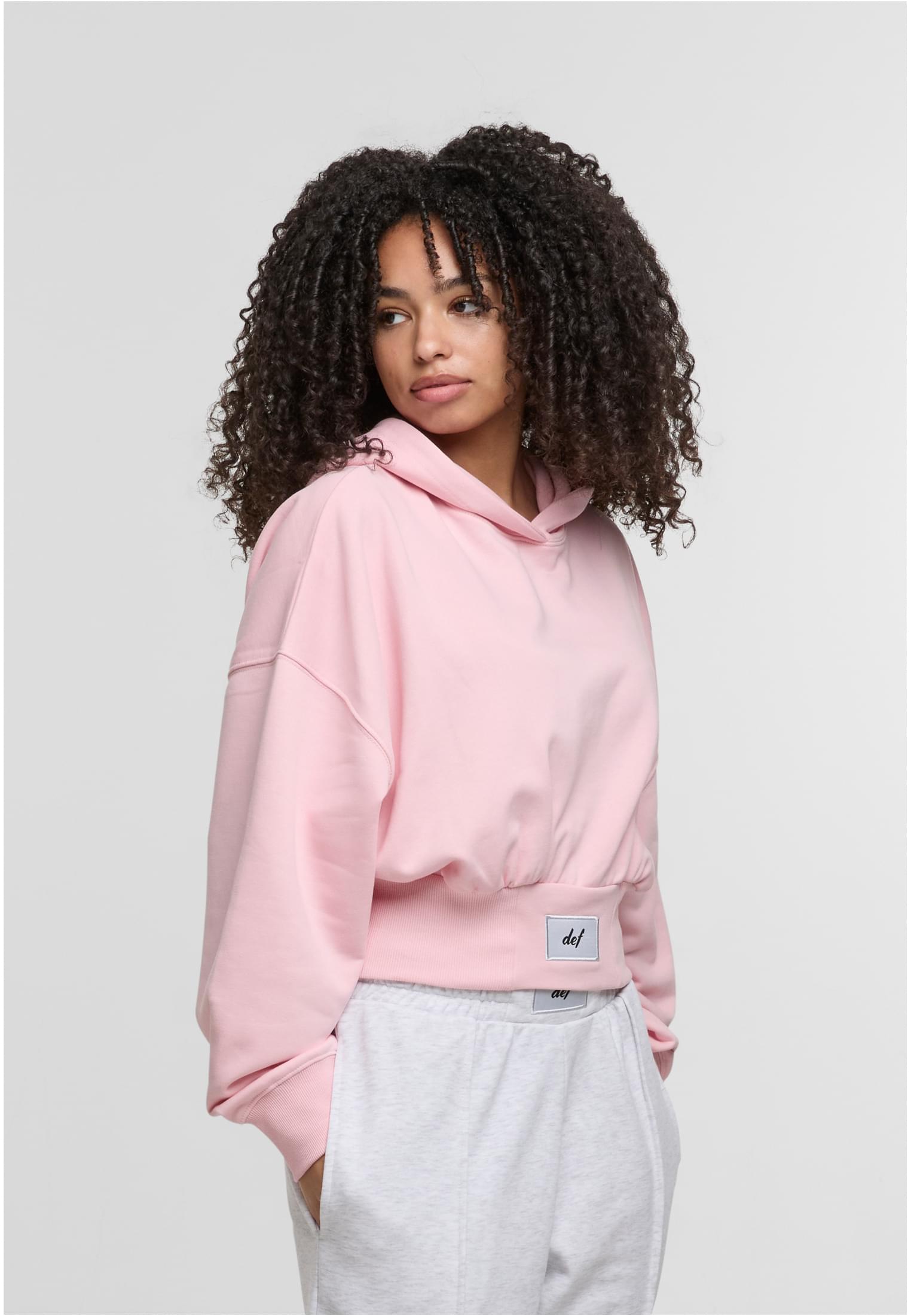 En kvinna med lockigt hår bär en ljusrosa kort DEF NICE Hoodie.