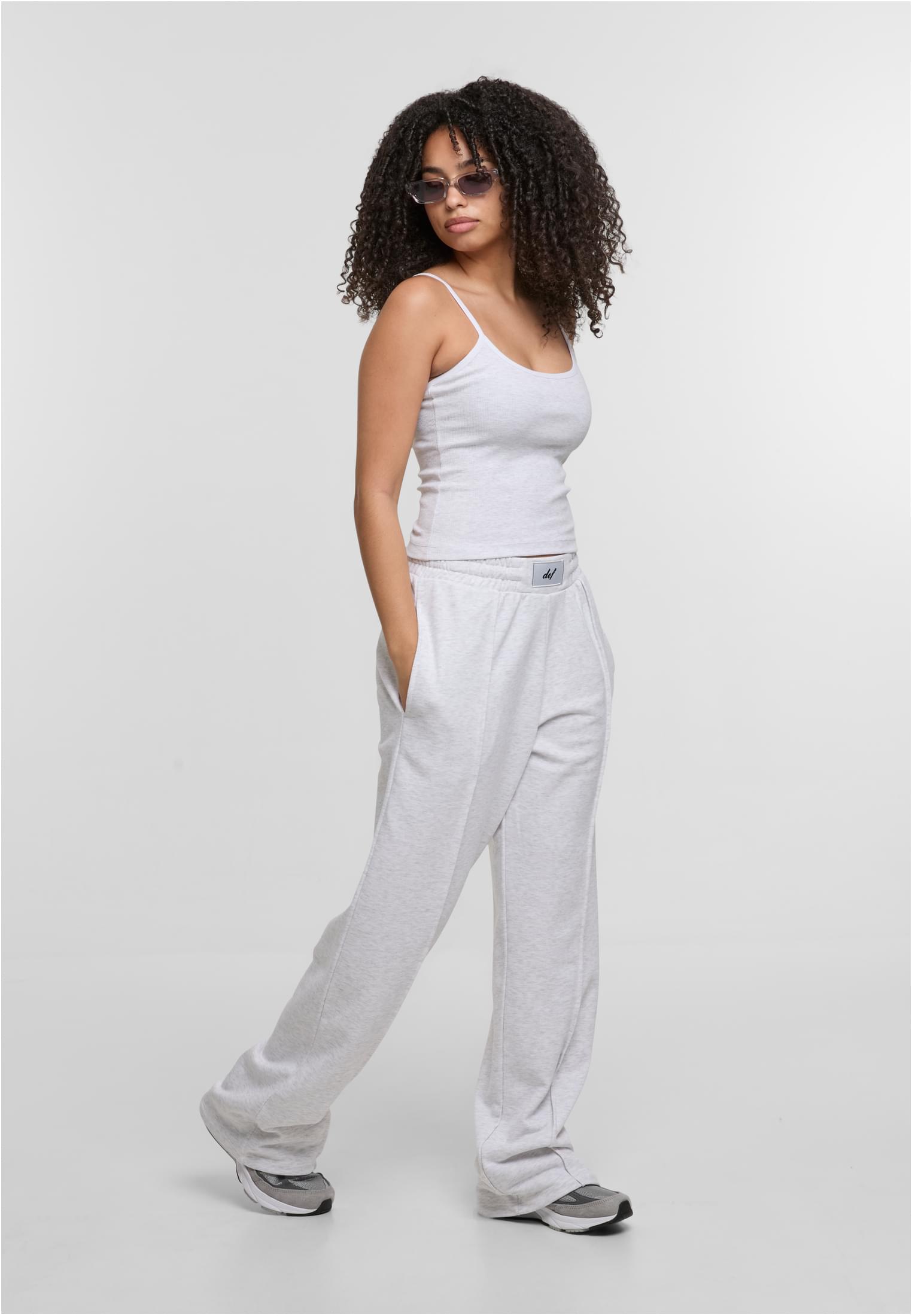 En kvinna med lockigt hår och solglasögon poserar från sidan, klädd i ett grått linne och matchande mjukisbyxor.