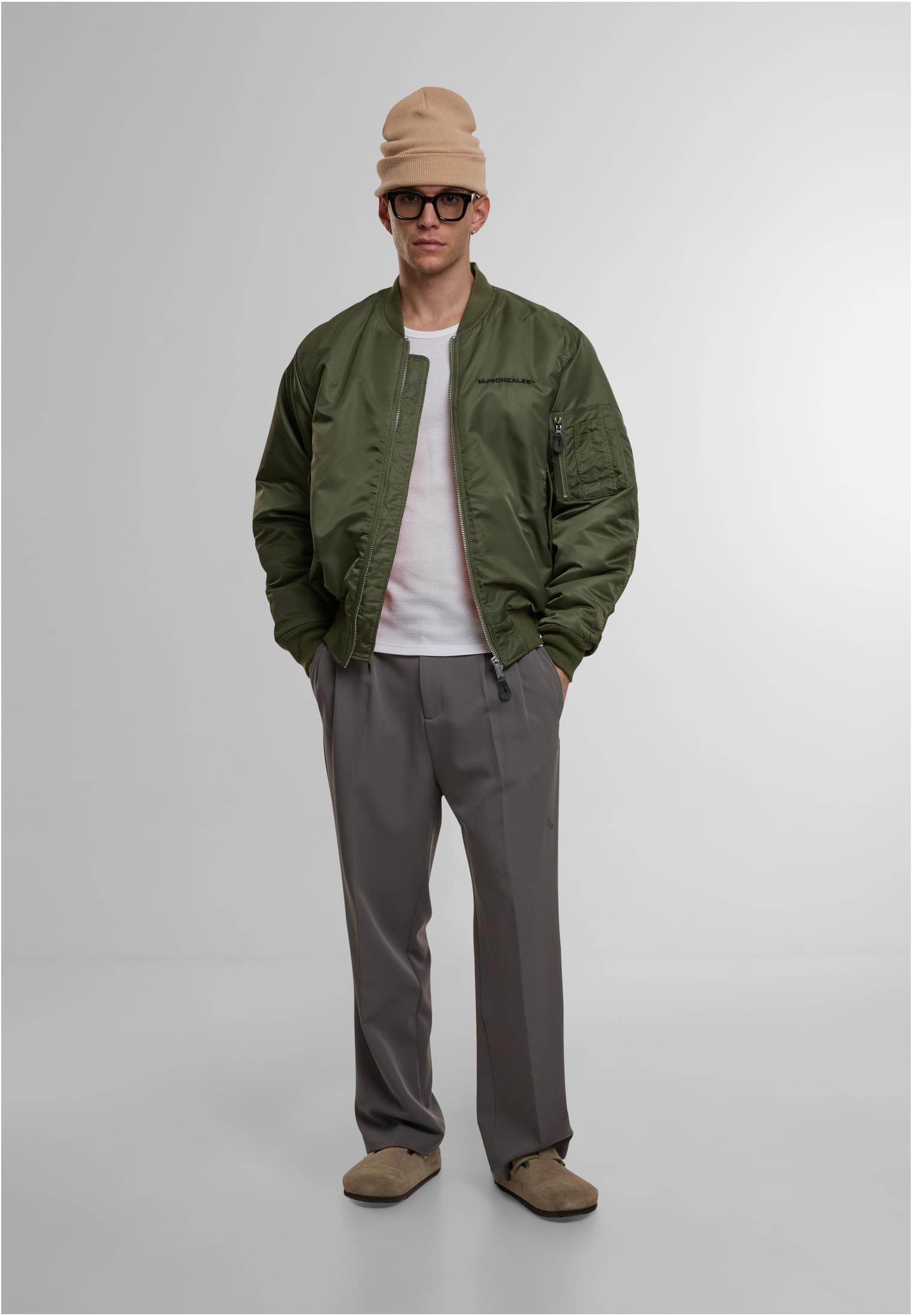 Framsida av en manlig modell som bär den olivgröna MJG Bomber Jackan, stylad med en vit t-shirt, grå byxor och en beige mössa.