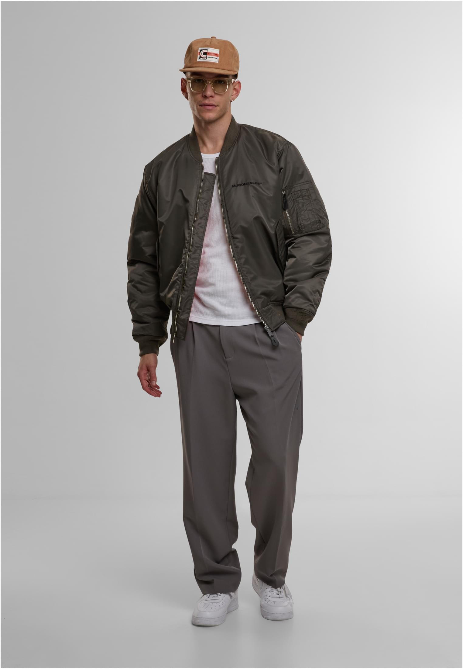 En manlig modell poserar i en olivgrön MJG bomberjacka, en vit t-shirt, grå byxor och en brun keps.