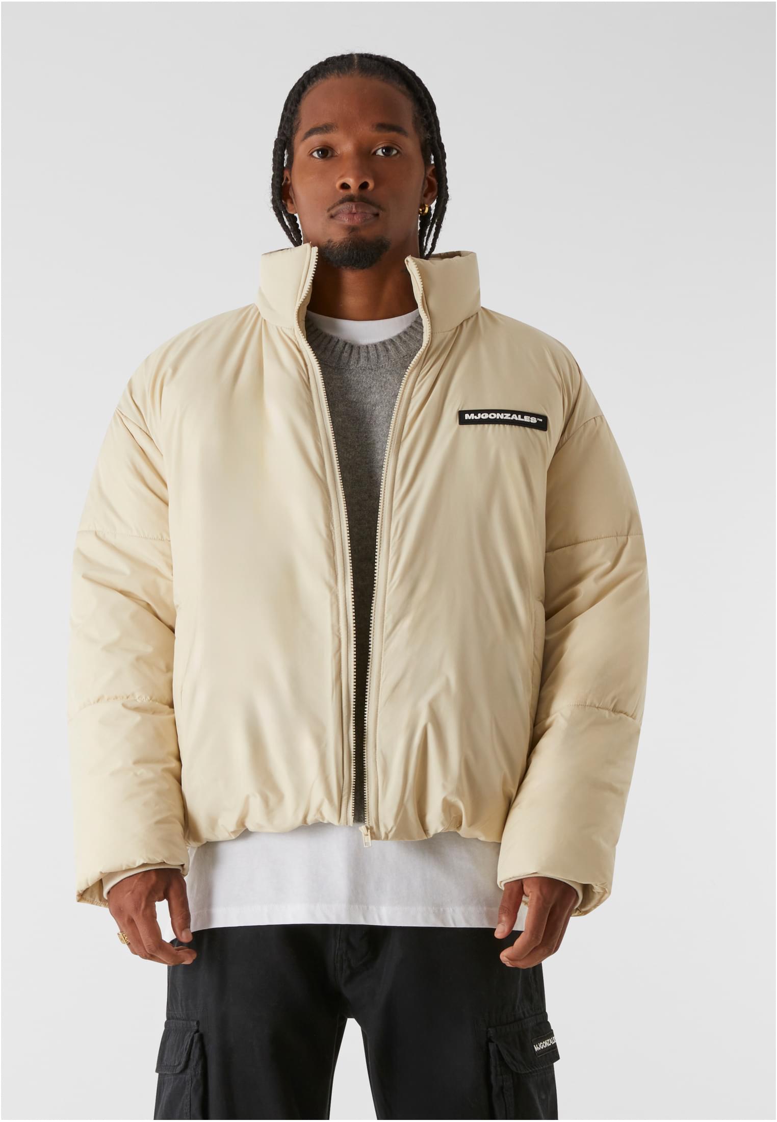 En man bär en öppen, beige MJG Puffer Jacket med en grå tröja och vit t-shirt under.