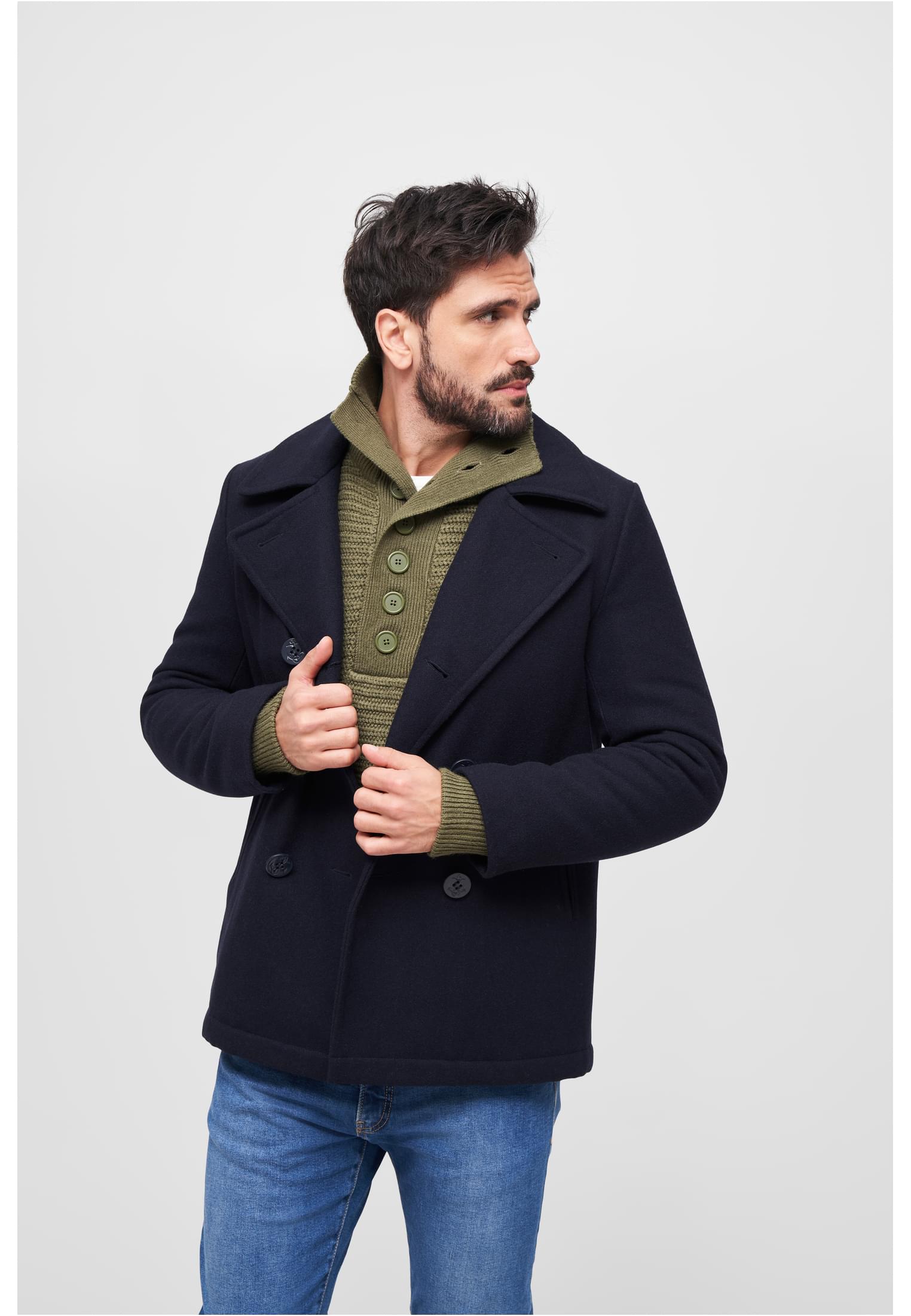 En man visar upp en mörk B Pea Coat skepparkavaj, stylad med en grön stickad kofta och jeans.