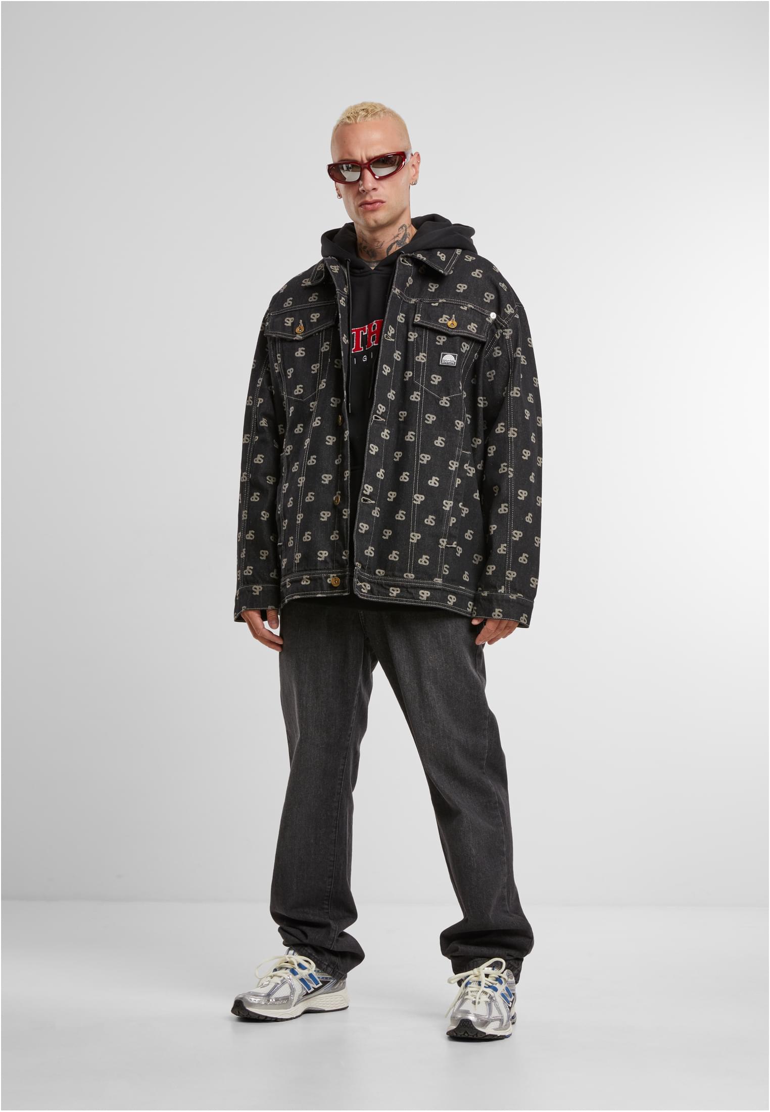 En man med blekt hår och solglasögon bär en svart jeansjacka med heltäckande mönster över en svart luvtröja och mörka jeans.