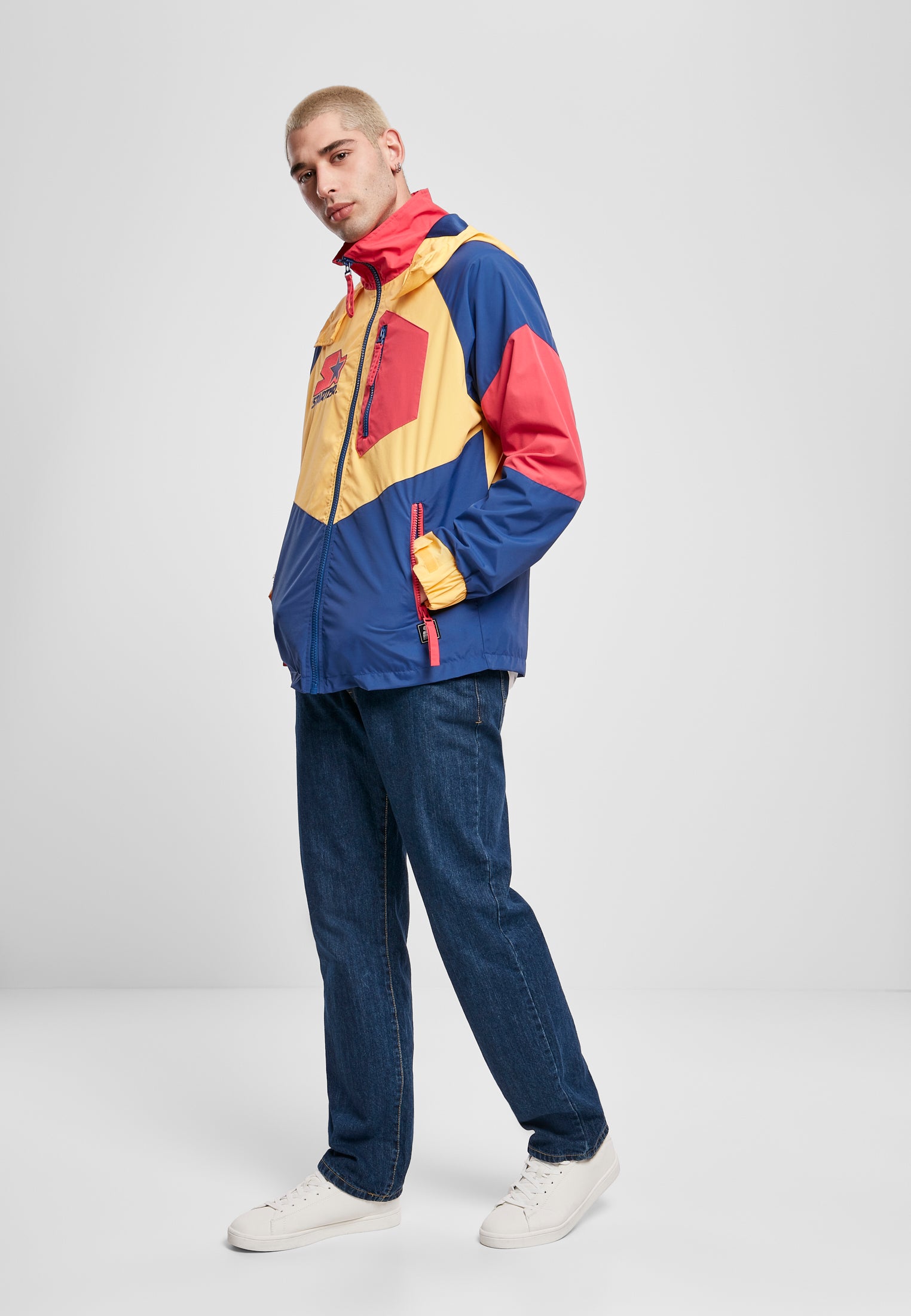 En man i trekvartsprofil som bär en SBL Starter Multicolored Logo Jacket i färgerna gult, blått och rött, tillsammans med blå jeans och vita sneakers.