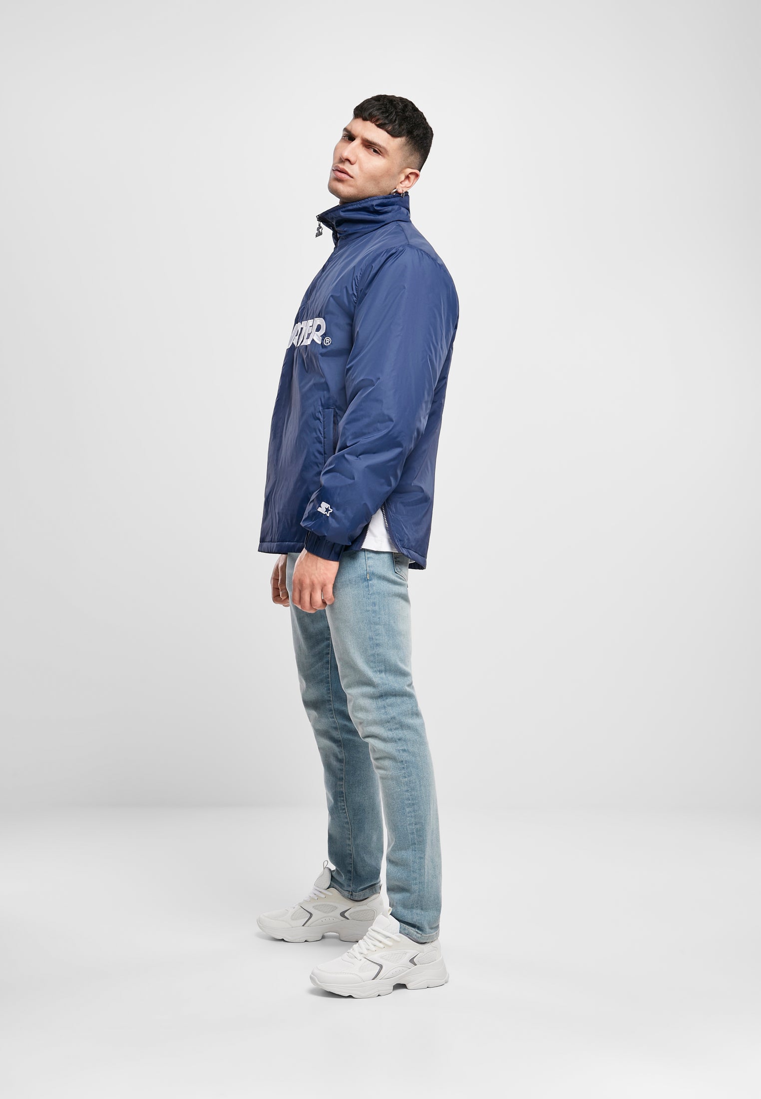 En man poserar från sidan i en blå SBL Starter Logo Half Zip Jacka, ljusa jeans och vita sneakers mot en vit bakgrund.