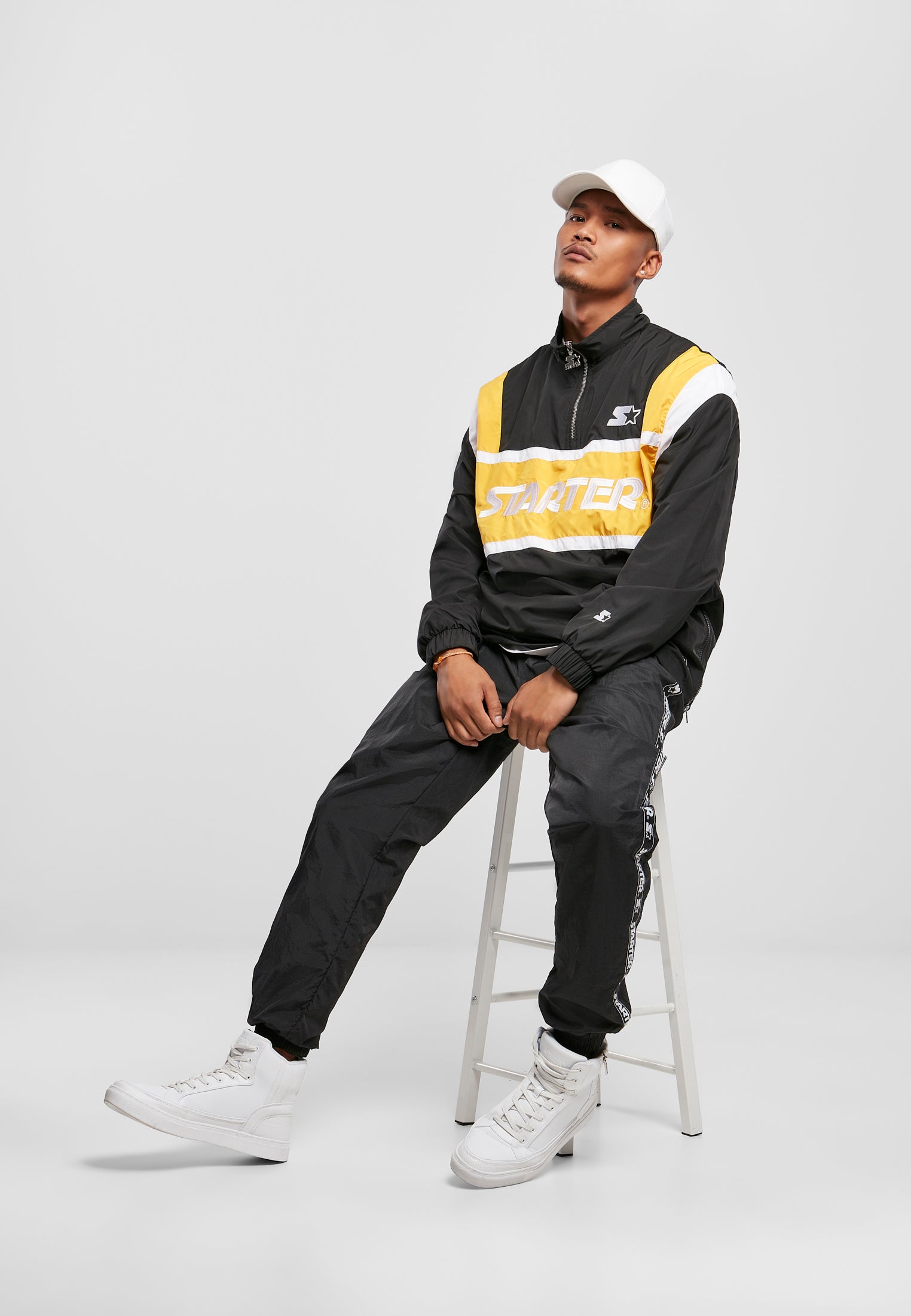 En man sitter på en vit pall iklädd en svart, gul och vit SBL Starter Half Zip Retro Jacka, svarta byxor och vita skor.