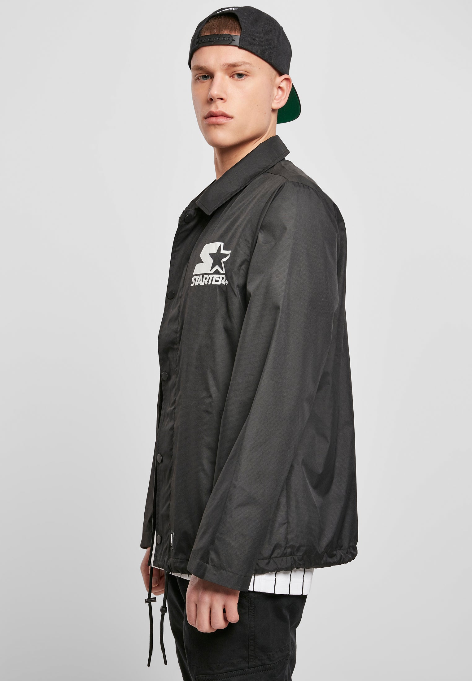 Sidovy av en man som bär en svart SBL Starter Coach Jacket med en vit logotyp på bröstet.