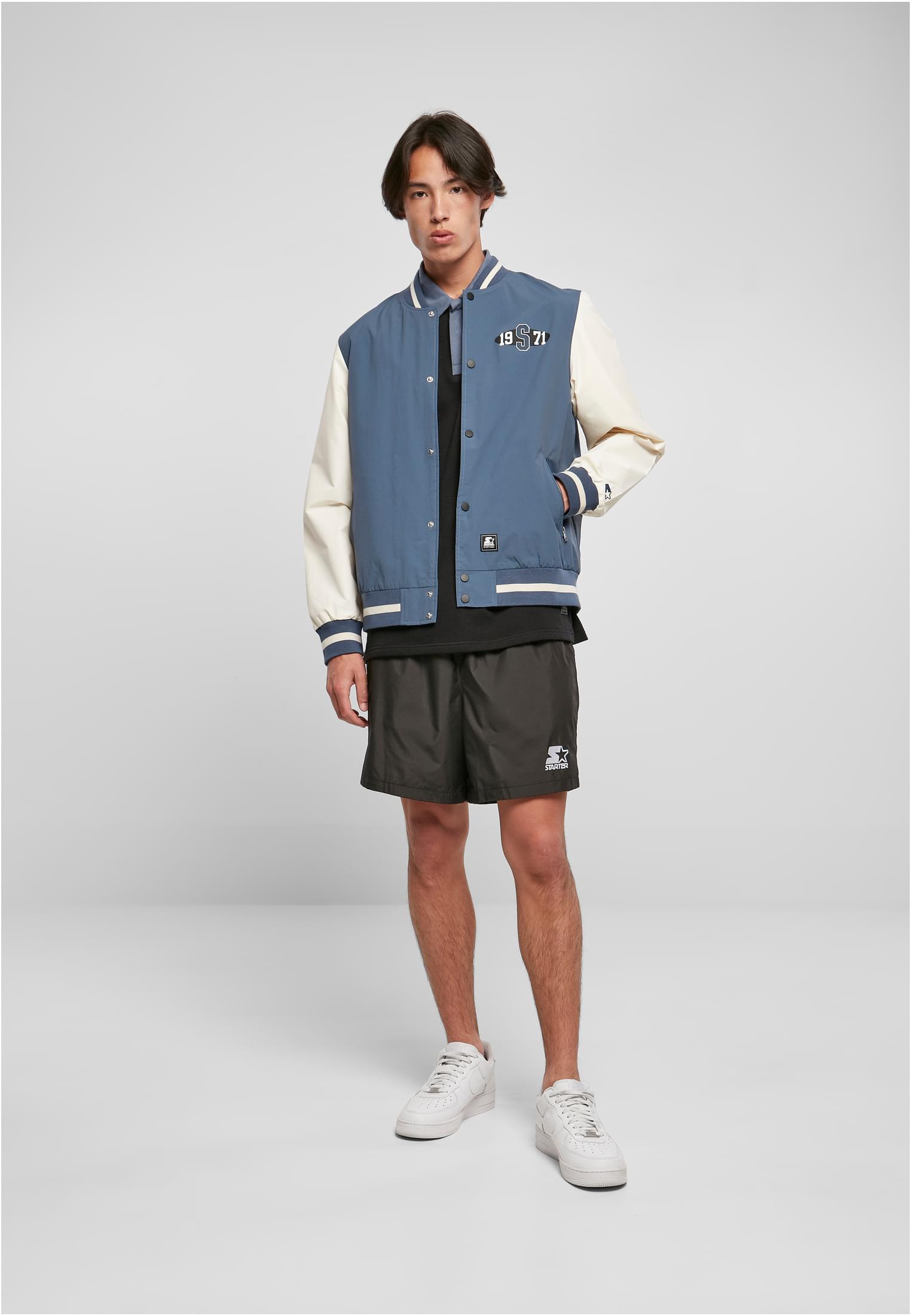 En man som bär den blå och vita SBL Starter Nylon College-jackan med svarta shorts, en svart t-shirt och vita sneakers.