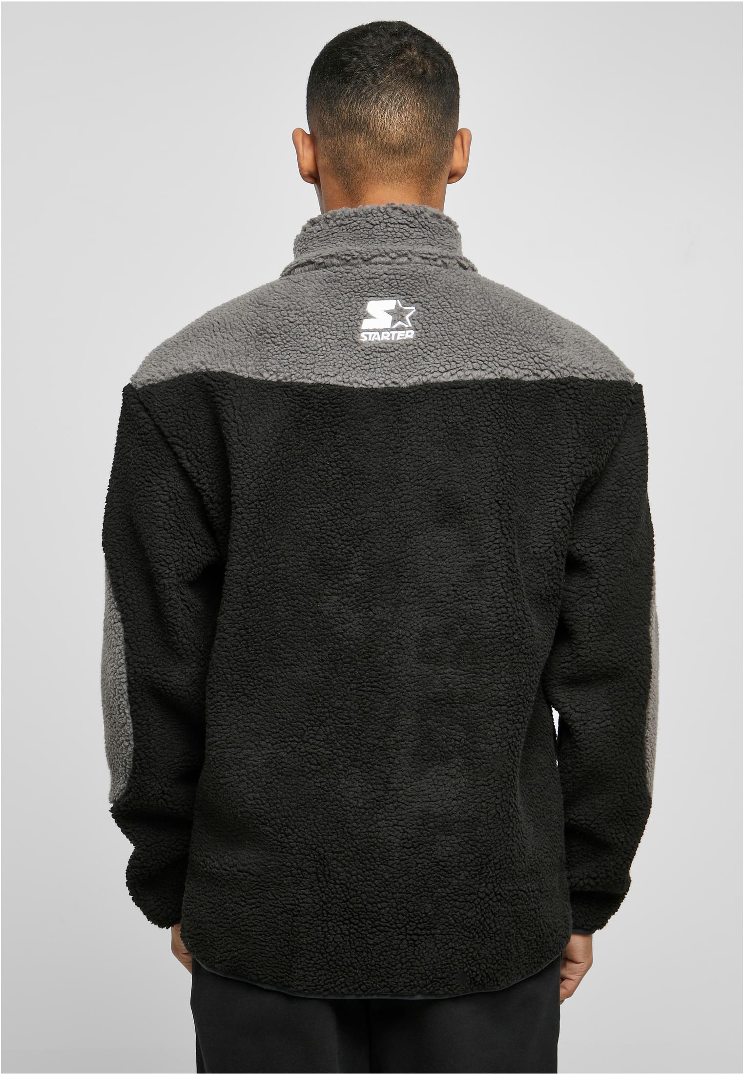 En man visar baksidan av en SBL Starter Sherpa Fleece Jacket i svart och grått, med en vit logga på ryggen.