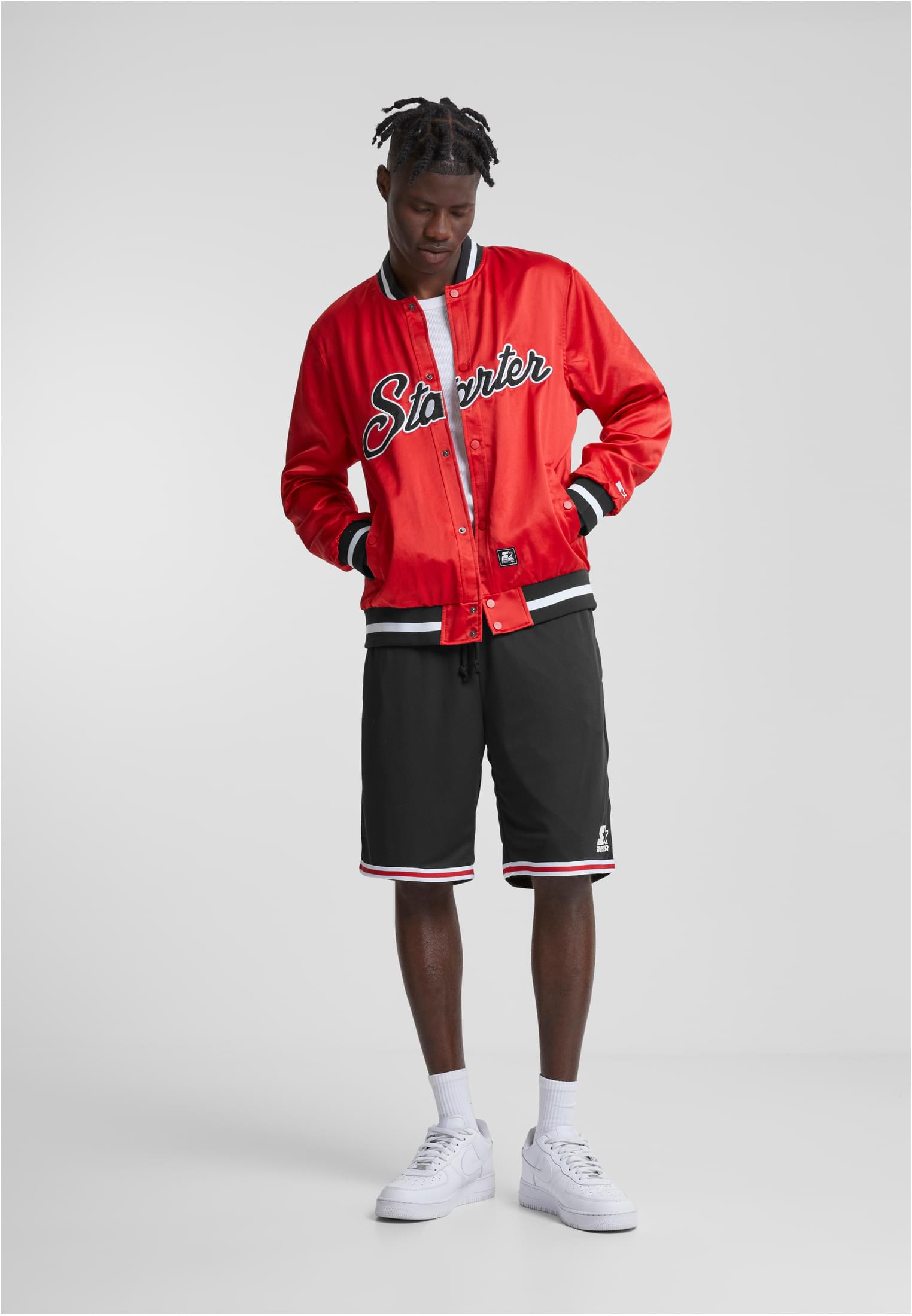En man visar framsidan av den röda SBL Starter Satin College Jackan, stylad med svarta shorts och vita sneakers.