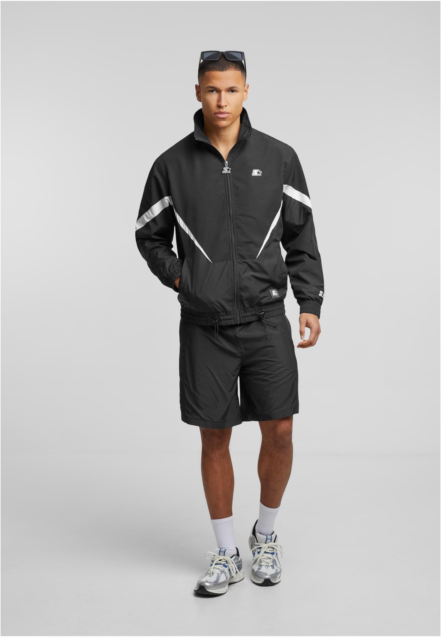 En man som bär en svart SBL Starter Fabric Mix Track Jacket med vita detaljer, tillsammans med matchande shorts och sneakers, fotograferad mot en ljusgrå bakgrund.