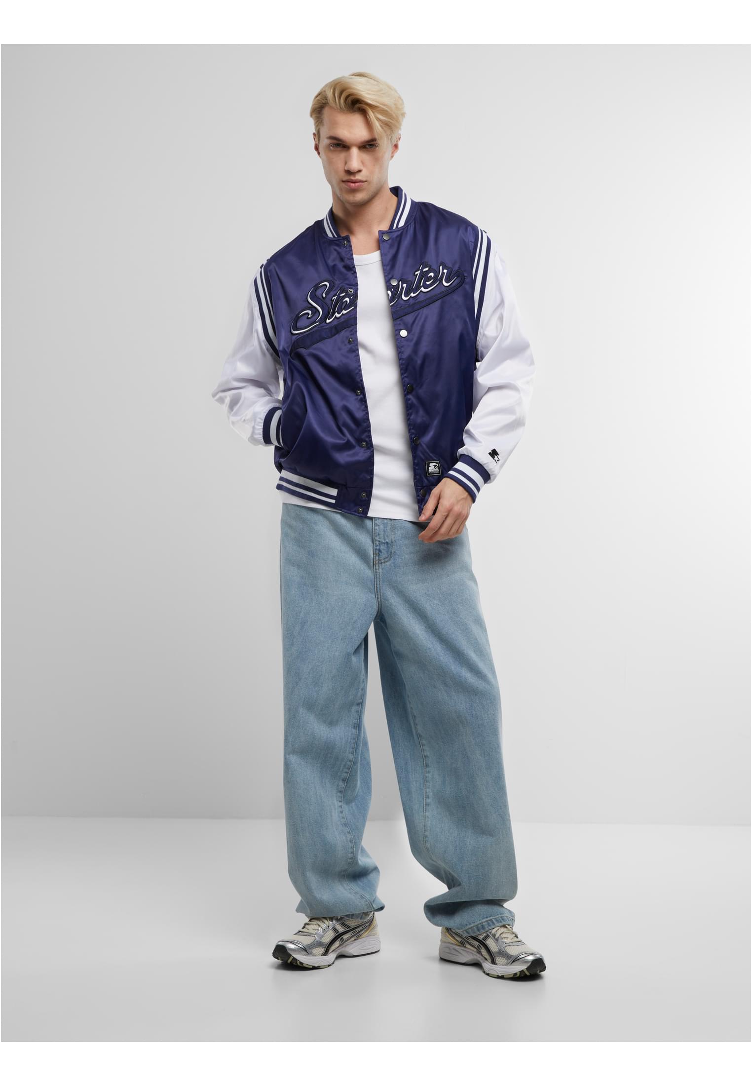 En manlig modell poserar i en marinblå och vit SBL Starter Satin College Jacket, stylad med en vit t-shirt och ljusa, vida jeans mot en vit bakgrund.
