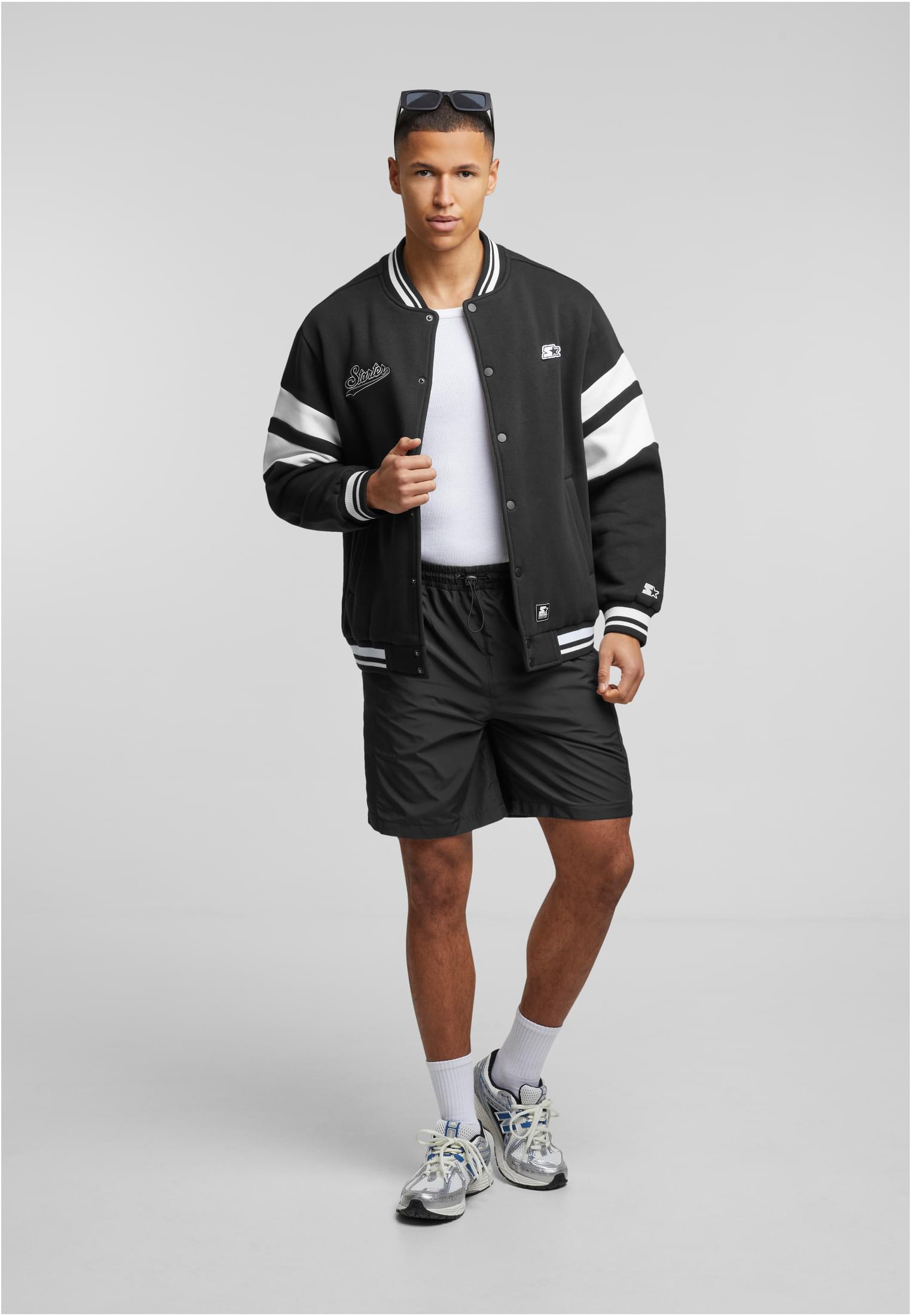 En manlig modell visar framsidan av en svart SBL Starter Sweat College Jacket, kombinerad med svarta shorts och en vit t-shirt.