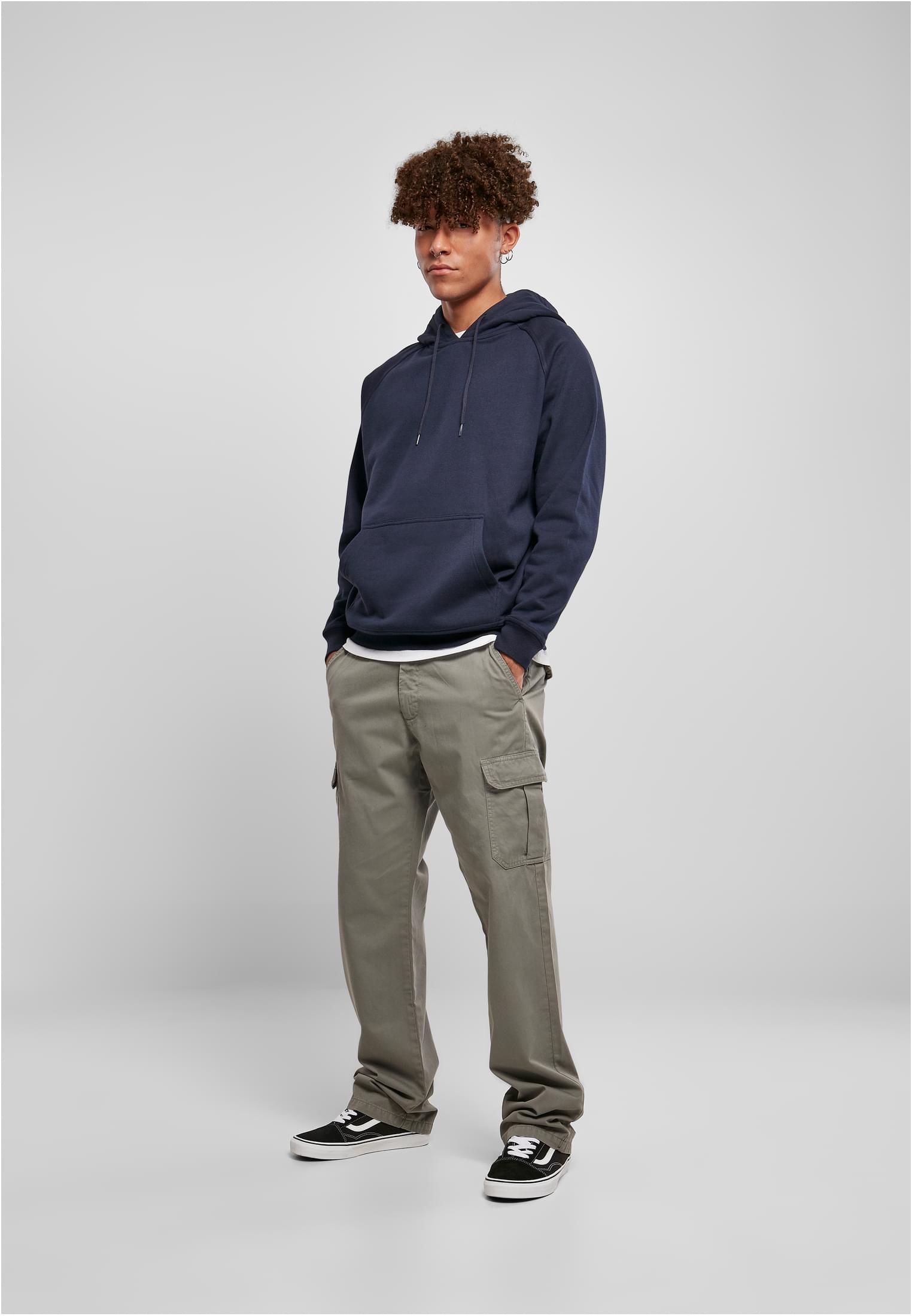 En man poserar i en mörkblå huvtröja, khakifärgade cargobyxor och svarta sneakers framför en ljusgrå bakgrund.