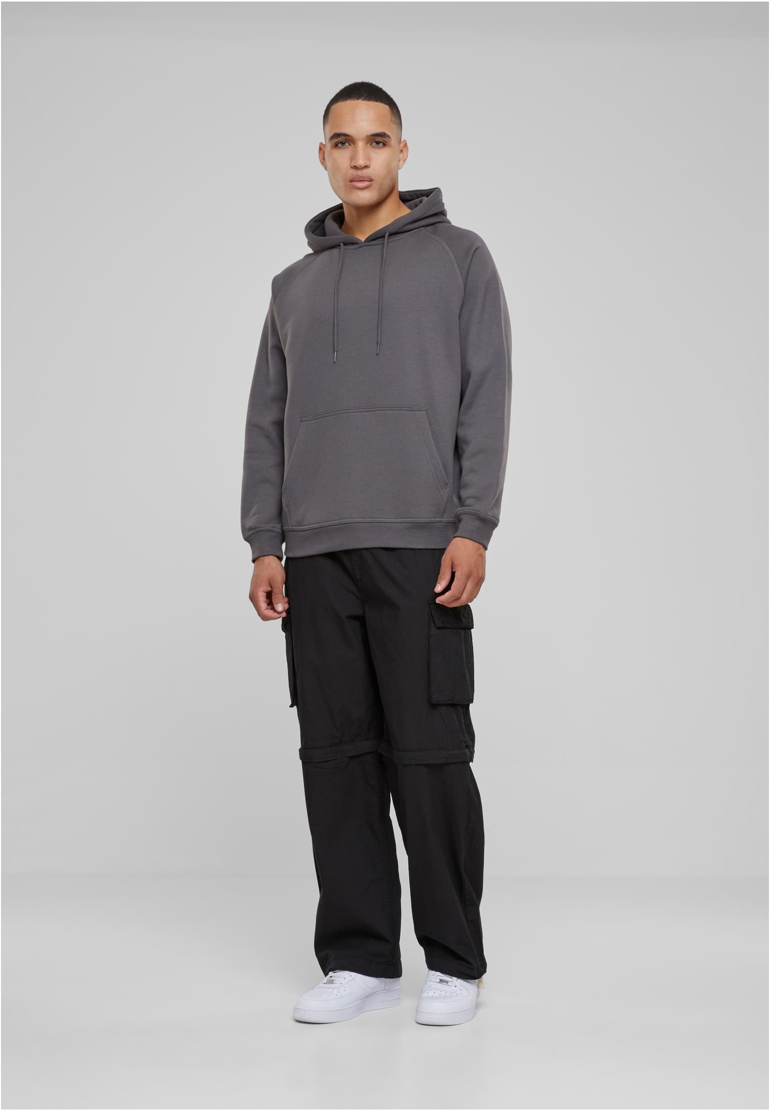 En man poserar framifrån i en mörkgrå huvtröja, svarta cargobyxor och vita sneakers.