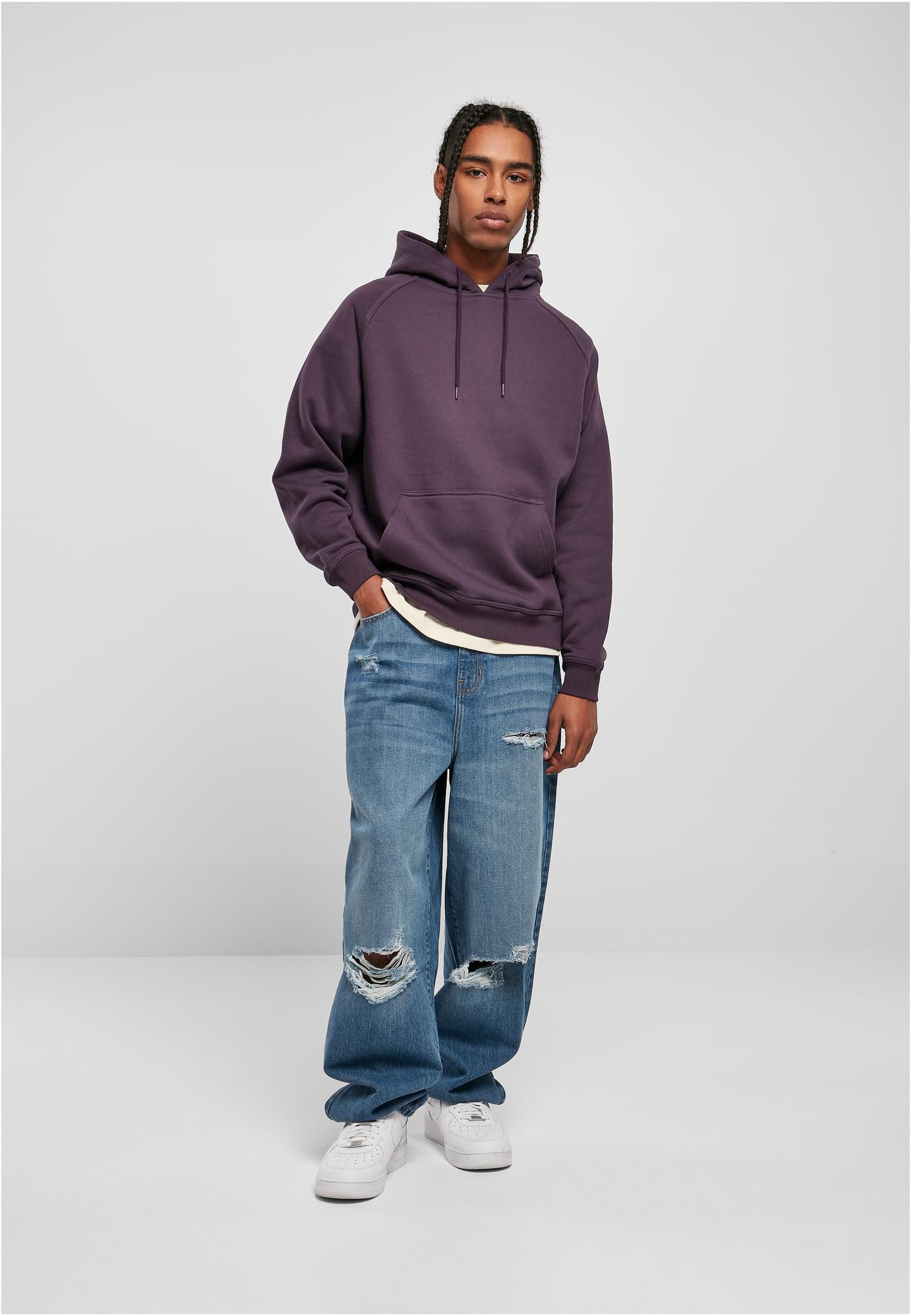 En manlig modell som bär en lila Urban Classics Blank Hoody med vida, slitna jeans och vita sneakers.
