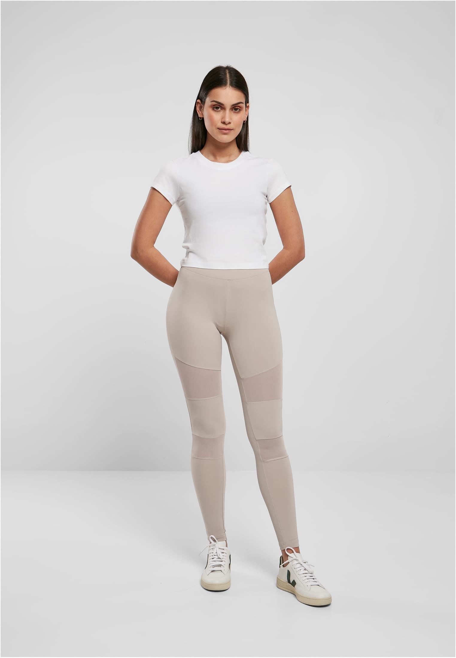 Framsida av en modell som bär Ladies Tech Mesh Leggings i warmgrey med mesh-paneler, tillsammans med en vit t-shirt och sneakers.