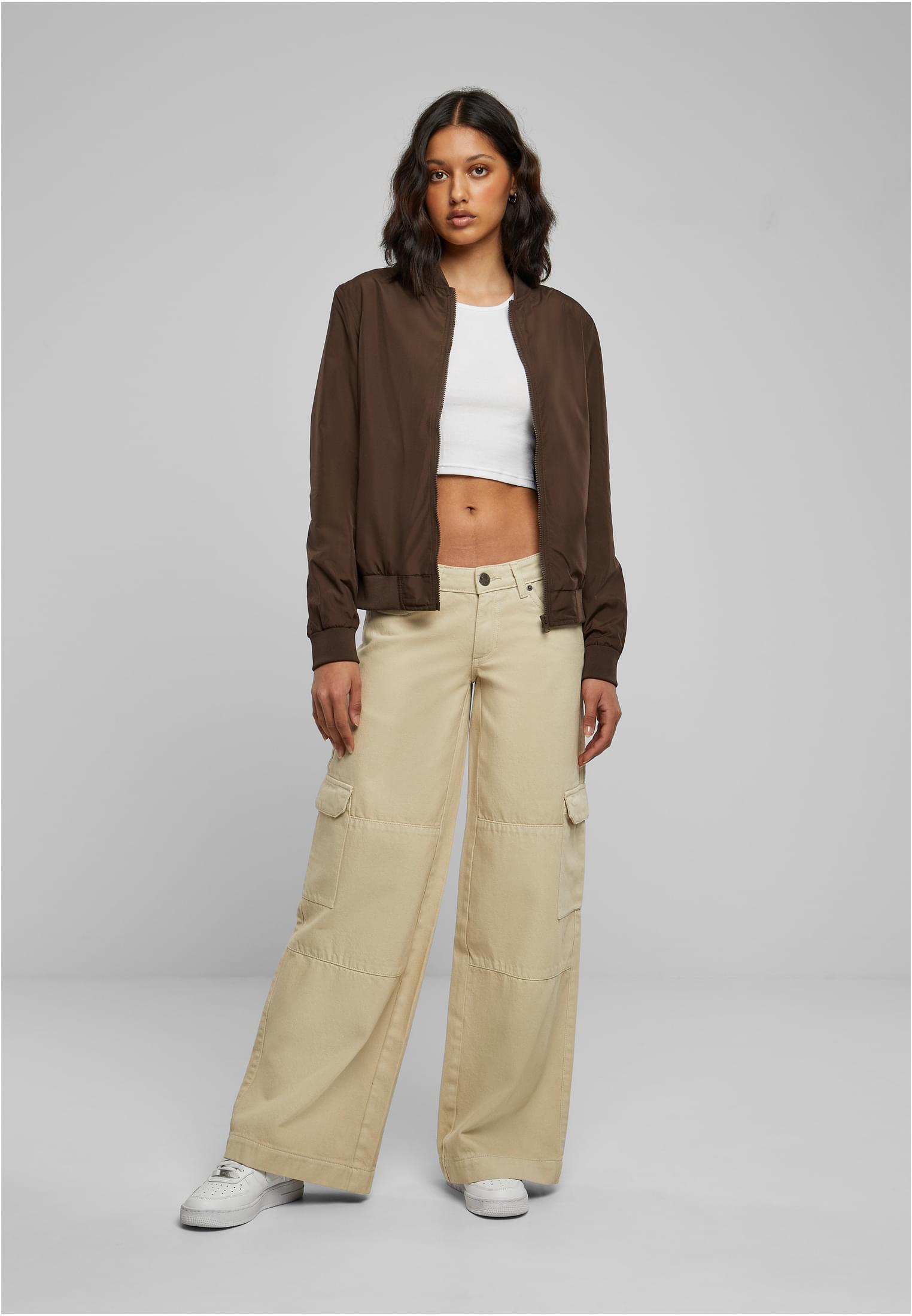 En kvinna visar framsidan av den bruna UC Light Bomber Jacket, stylad med en vit magtröja och beiga cargobyxor.
