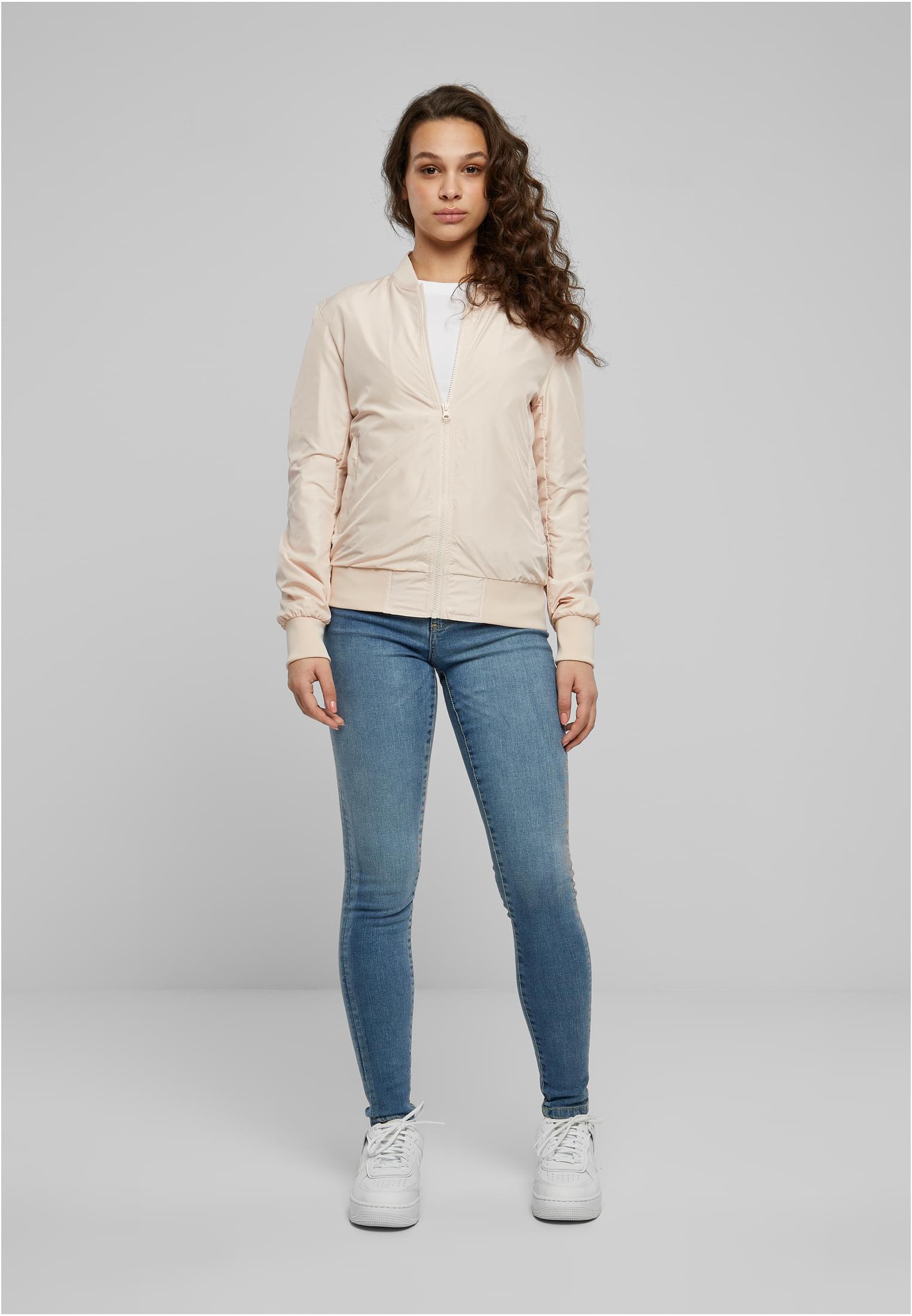 En kvinna bär en ljusrosa bomberjacka, blå jeans och vita sneakers.