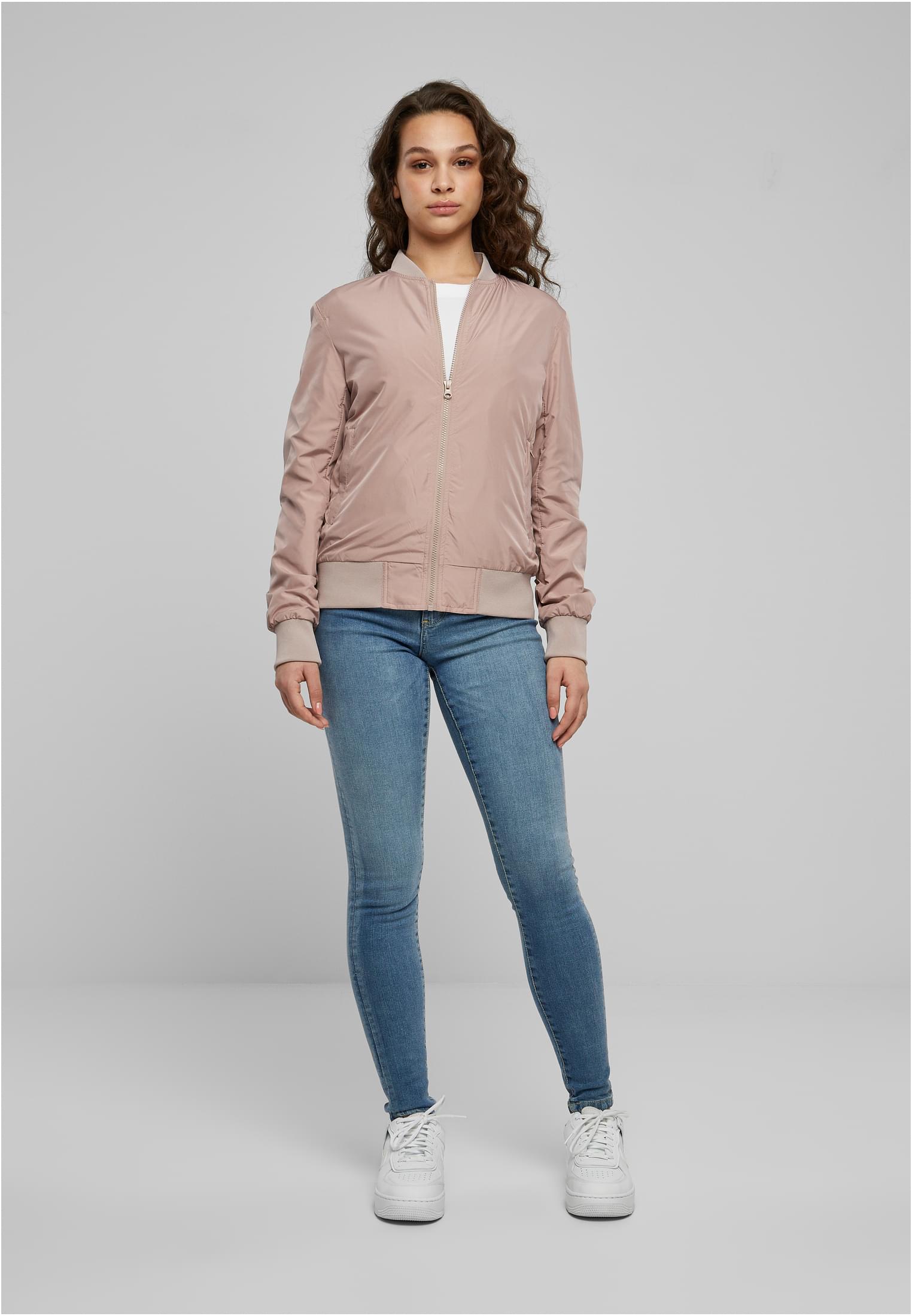 En kvinna visar framsidan av en ljusrosa UC Light Bomber Jacket, buren med blå jeans och vita sneakers.