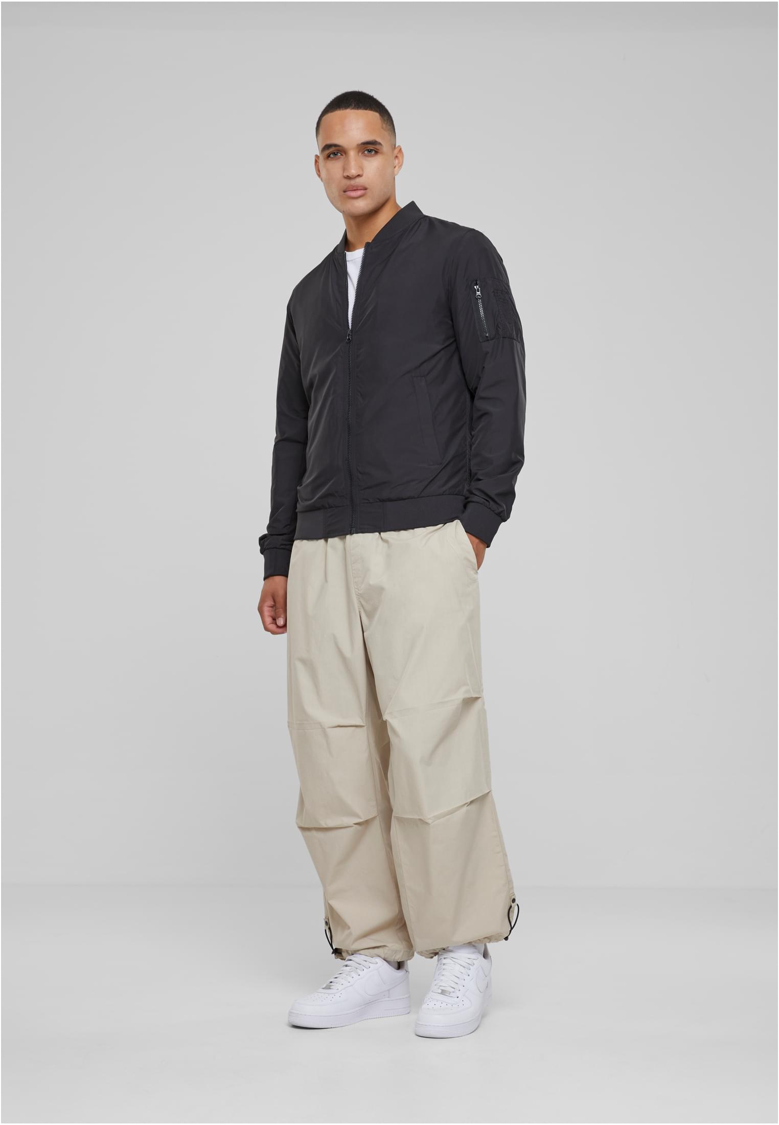 En manlig modell som bär en svart bomberjacka, vida beigefärgade byxor och vita sneakers.