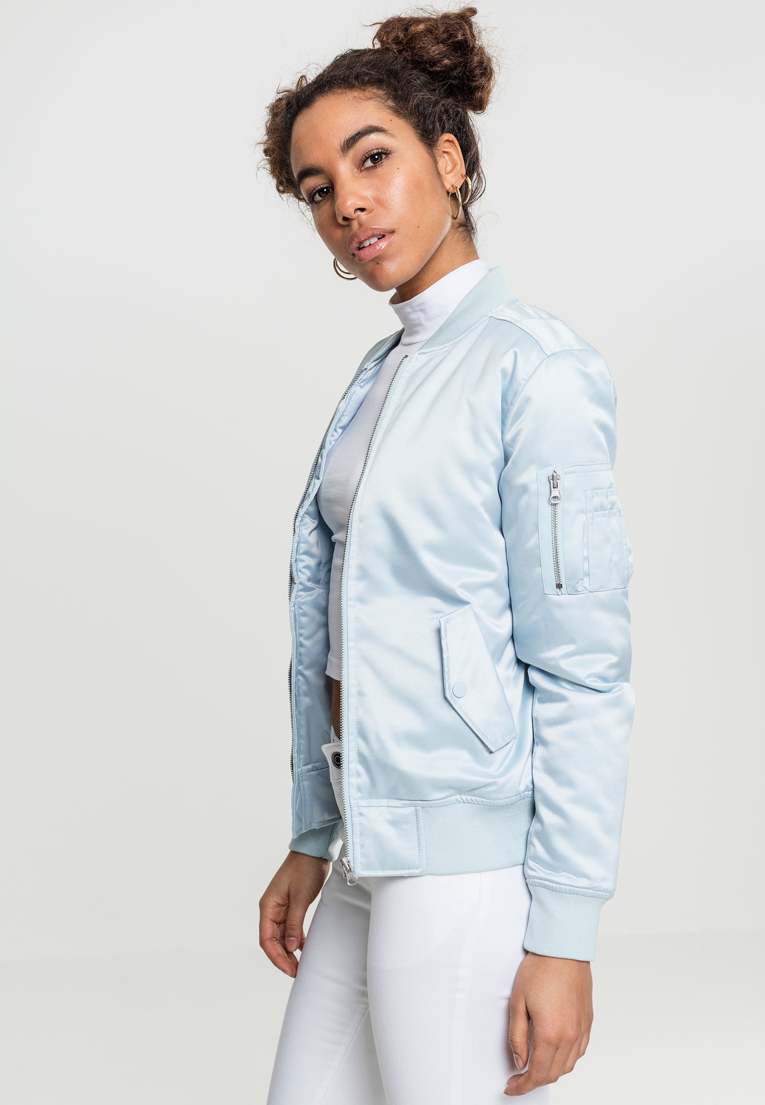 En kvinna bär en ljusblå UC Satin Bomber Jacket med vita byxor, fotograferad snett framifrån mot en ljus bakgrund.
