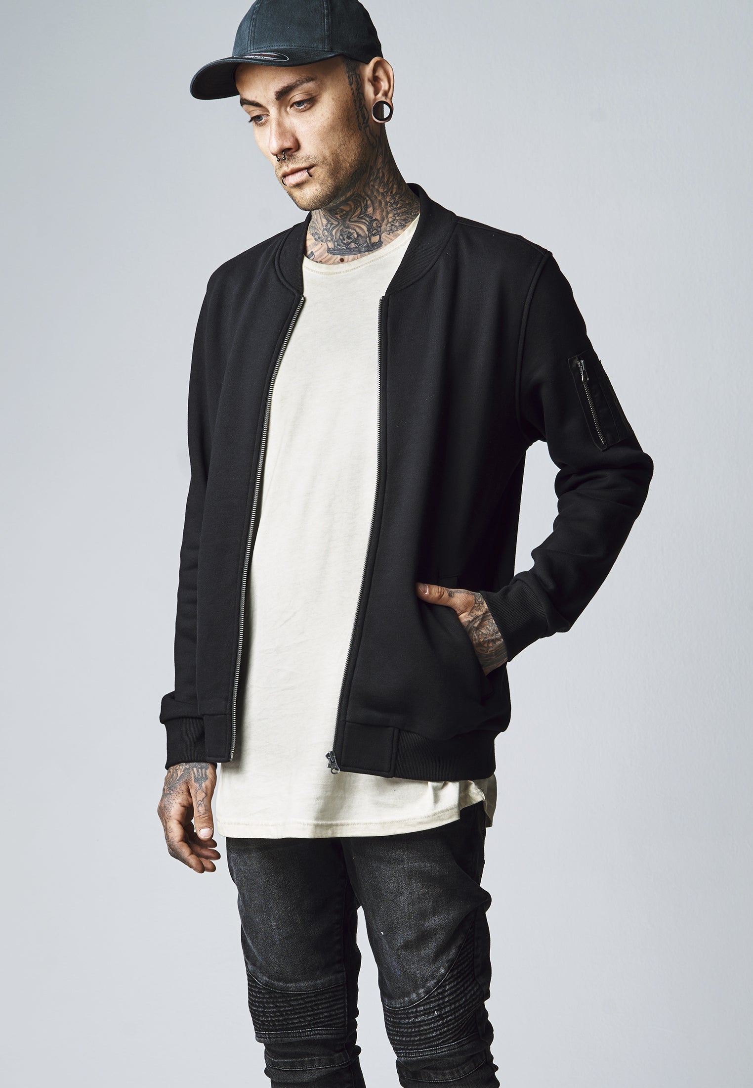 En man med tatueringar bär den svarta UC Sweat Bomber Jacket med handen i fickan.